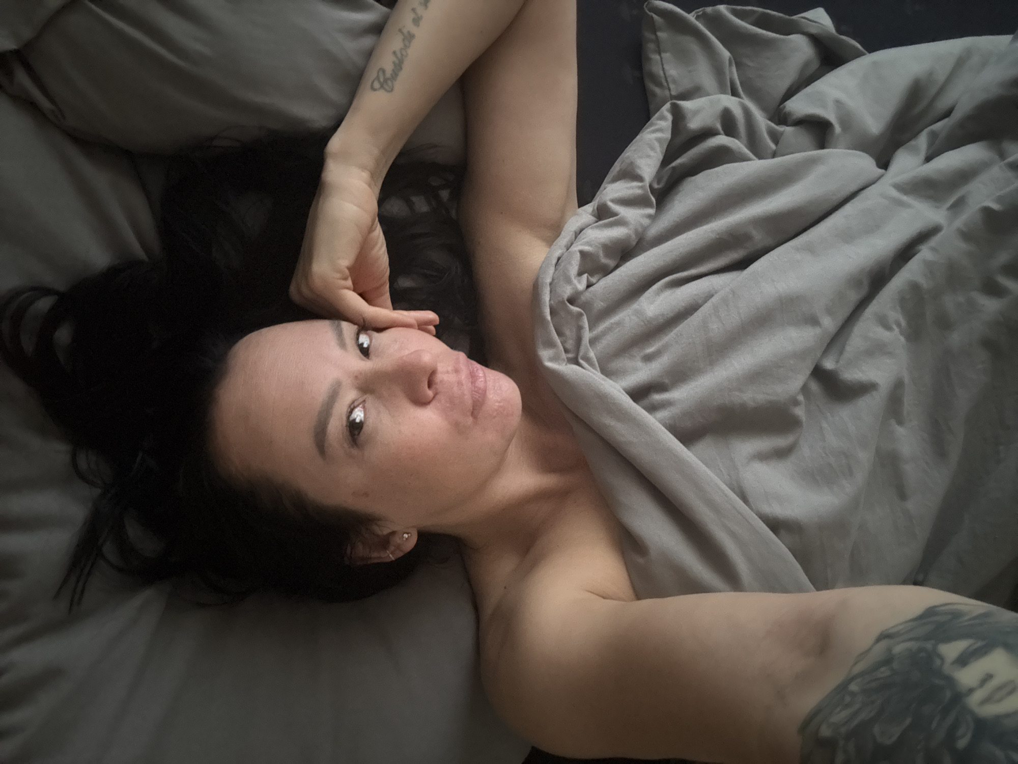 A woman is lying in a bed with gray sheets and a duvet. She has long, dark hair and is looking at the camera. Her left arm is raised with her hand near her face, and her right arm is extended downwards. Tattoos are visible on both arms. The location is not discernable from the image.