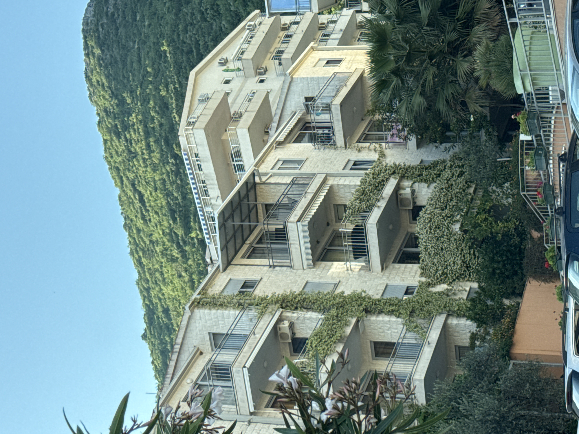 The image presents a multi-story building in what is presumed to be Nerin, Montenegro, based on the prompt. The architectural style is modern, featuring balconies with metal railings. The facade is light-colored, possibly stone or stucco, and is partially covered with climbing vegetation, suggesting a deliberate integration of nature into the structure. The building appears to be residential. There are no people visible in the scene. A heavily forested hillside forms the backdrop on one side. The foreground includes partial views of foliage, indicating the presence of landscaping around the building. The sky is clear and blue. A portion of a vehicle is visible in the right of the frame.