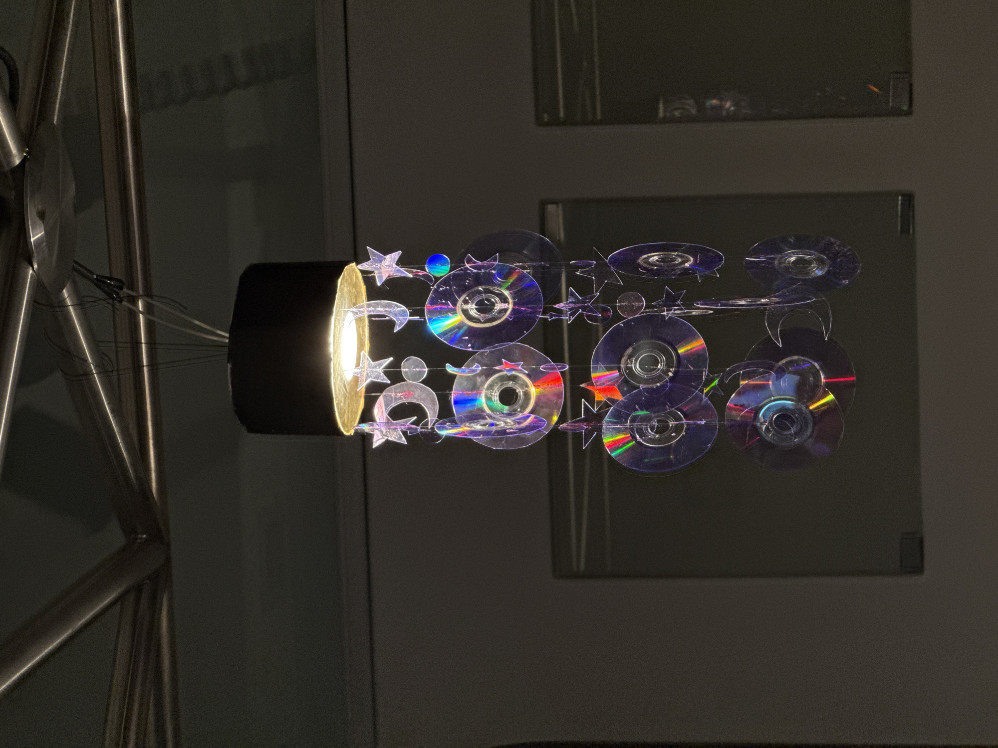 A close-up shot shows a self-made light fixture. The fixture consists of a black cylinder with a glowing golden-yellow light emanating from one end. Adorning the emitted light are several translucent or transparent objects, including stars, moons, and round discs that appear to be CDs. These objects are suspended in a way that makes them appear to be emanating from the light source. The light fixture is attached to a metallic structure of rounded tubes. In the background are dark walls with rectangular patches that appear to be framed photos or paintings. The scene is dimly lit, focusing the attention on the light fixture itself. The location is not possible to determine from the given image and cannot be provided.