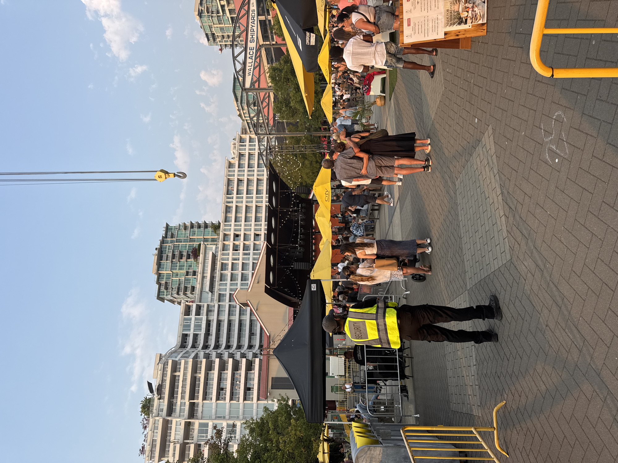 This image depicts an outdoor event at Wallace Shipyards in North Vancouver, Canada. In the foreground, a security guard wearing a yellow reflective vest and black pants stands facing away from the camera. Behind him, a crowd of people is gathered, some standing and some sitting at tables beneath yellow and black umbrellas. A stage with lighting is visible in the background, suggesting a performance or event is taking place. Several modern apartment buildings and industrial structures, including a large crane, are present in the background, indicating an urban waterfront setting. The ground is paved with brick-like stones. The lighting suggests it is late afternoon or early evening.
