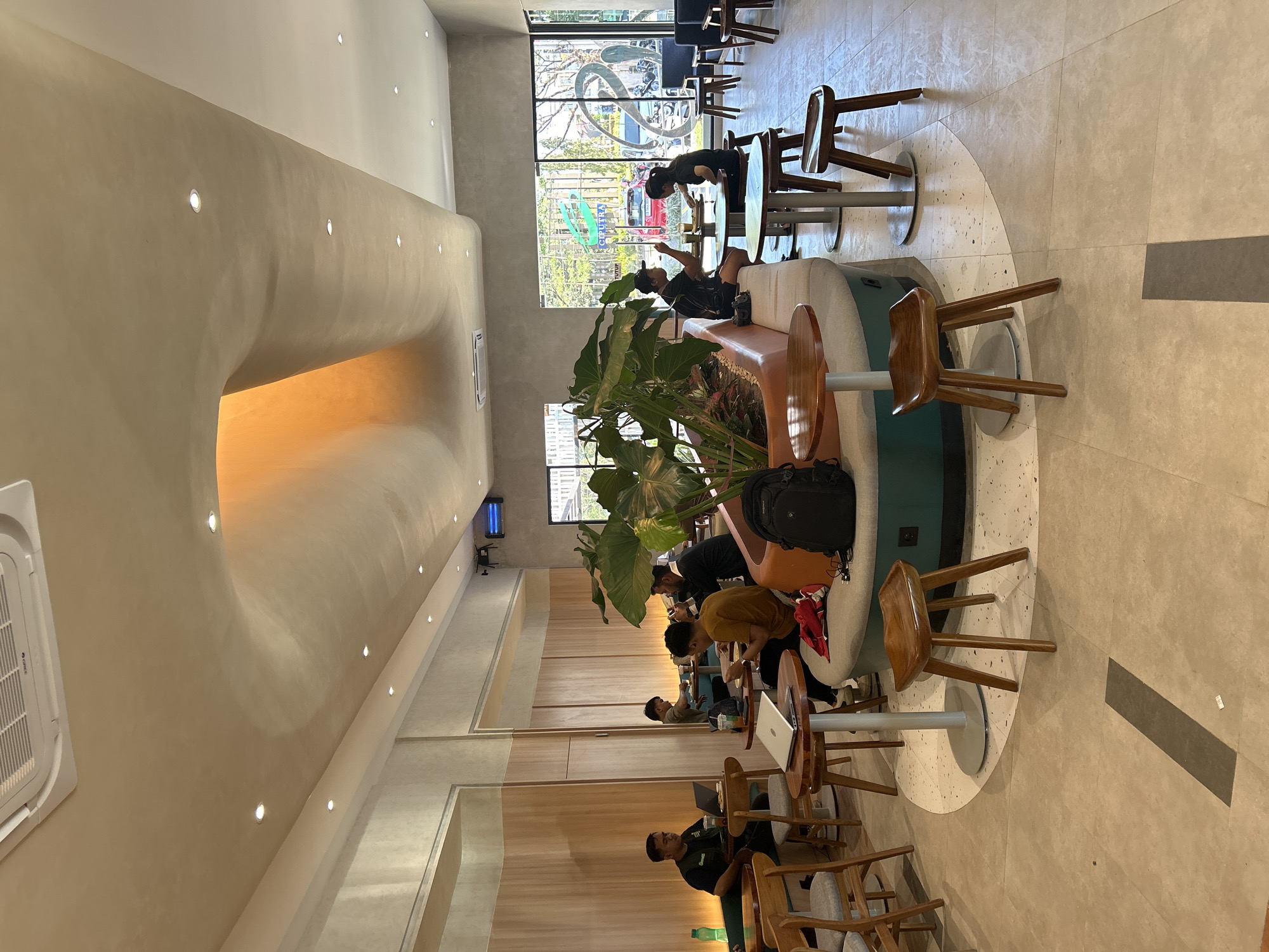 The image depicts the interior of a modern coffee shop, possibly located in Tombolo, Indonesia, characterized by its contemporary design and relaxed atmosphere. The space features a curved ceiling with recessed lighting, creating a warm ambiance. A large, central planter filled with lush greenery serves as a focal point, surrounded by a semi-circular seating area with plush upholstery. Various tables and chairs, made of wood and metal, are arranged throughout the coffee shop, accommodating patrons.
Several individuals are present, engaged in diverse activities. In the foreground, a person is seated at a round wooden table, possibly working on a laptop. To the left, another individual is also seated at a table, seemingly engrossed in their work. Further back, a group of people are gathered around tables, some interacting with each other, while others are focused on their devices. The overall impression is that of a busy yet orderly coffee shop, where patrons can socialize, work, or simply unwind.
The decor is minimalist, with a combination of wood paneling on the walls and neutral-toned flooring. Large windows offer a glimpse of the outdoors, suggesting the coffee shop is situated in an urban environment. The presence of plants adds a touch of nature to the indoor setting, enhancing the overall aesthetic appeal. The image captures a slice of everyday life in what appears to be a popular and well-designed coffee shop.