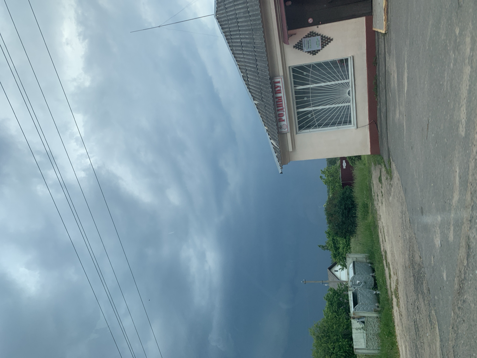 The image shows a roadside scene with a small building and a stormy sky. The building, which appears to be a shop or kiosk, has a light-colored facade with a darker trim around the base and a corrugated roof. A sign above the window reads "РОДНЫ КУТ" in red lettering. The window is covered with a decorative white metal grate. To the left of the building, there are two large metal trash bins. In the background, there are trees, a fence, and a house. The sky is dark and overcast with visible storm clouds. The overall impression is a rural or semi-rural setting in Слабада-Кучынскі сельскі Савет, Belarus, under threatening weather conditions.