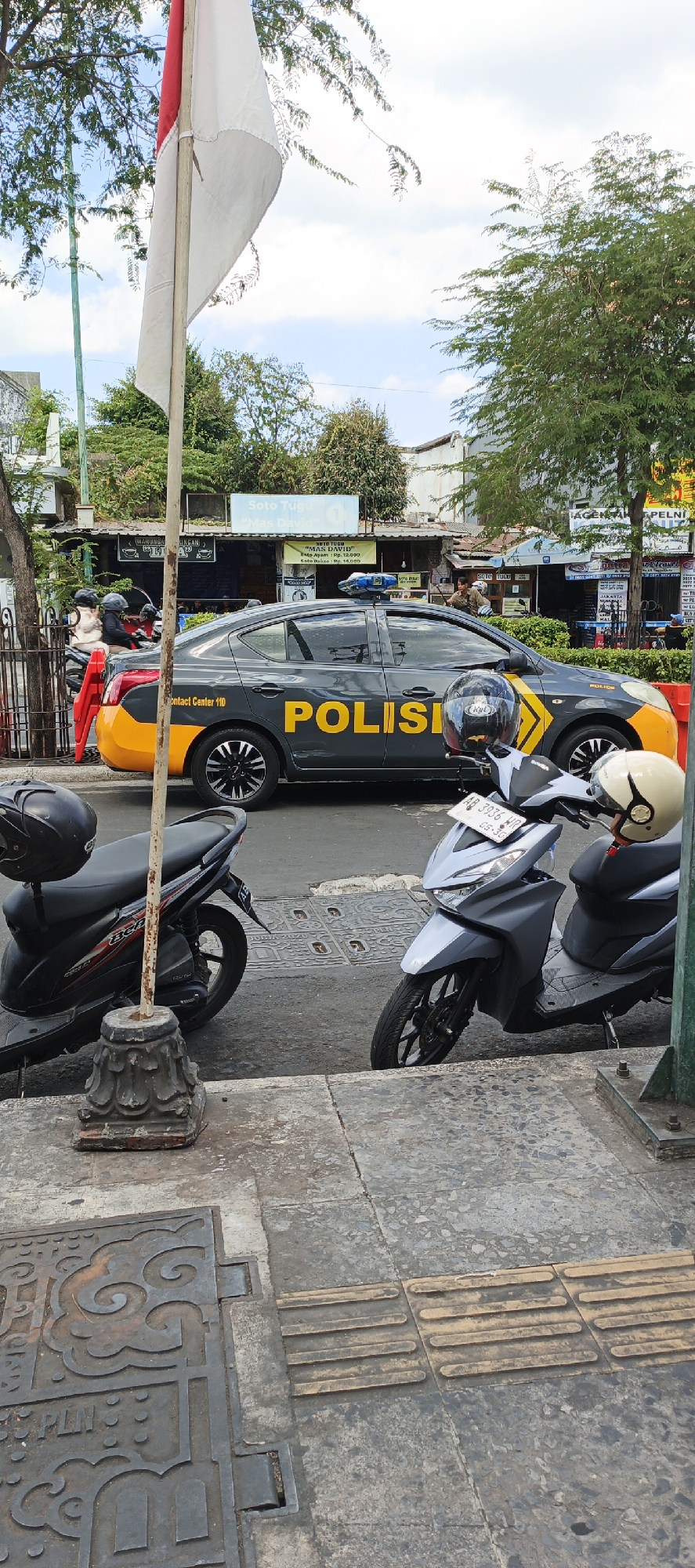 The image captures a street scene in Yogyakarta, Indonesia, with a police car and several motorcycles parked on the roadside. The police car, marked with "POLISI" in large yellow letters, is dark gray with yellow accents on the rear. It has a light bar on its roof, indicating it's an official vehicle. Two motorcycles are parked in the foreground. The one on the left is black with red accents, and the one on the right is light gray. Both have helmets on them.
Behind the police car, there are several small shops with signs. One sign reads "Soto Tugu 'Mas David'". People are visible walking and sitting in the background, suggesting a commercial area. The street is paved, and there's a sidewalk in the foreground with tactile paving for the visually impaired. A flagpole with the Indonesian flag is also prominent in the left part of the frame. The sky is visible above, with scattered clouds. The overall impression is a busy urban environment with law enforcement presence.