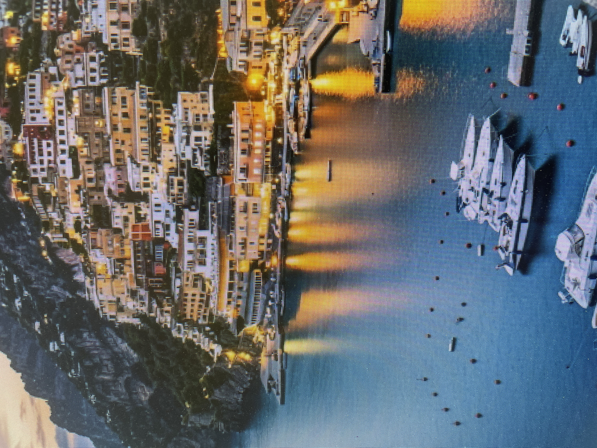 The media file displays a vibrant, densely built coastal town nestled into a dark, rocky hillside, illuminated at dusk or night. The image is oriented with the town on the left and a harbor on the right. Numerous multi-story buildings, characterized by their light-colored facades and numerous windows, cascade down the slopes, each window glowing with warm, artificial light. These lights cast long, golden reflections onto the calm, dark blue waters of the harbor.
The harbor is filled with various boats, including several large white yachts and smaller vessels, some appearing to be docked or moored. Small red buoys are scattered across the water. The overall scene suggests a picturesque and bustling port city. No specific human activity is discernable, but the lit windows imply life within the buildings. The image has a visible pixelated quality, indicating it might be a photograph of a screen or a low-resolution digital capture.
Based on the visual evidence, which clearly shows a mountainous coastal settlement with a harbor full of boats and distinctive architecture often found in Mediterranean regions, the scene does not align with the provided location context of Poltava, Ukraine, which is an inland city without such coastal features or mountainous terrain. There is no visible text in the image.