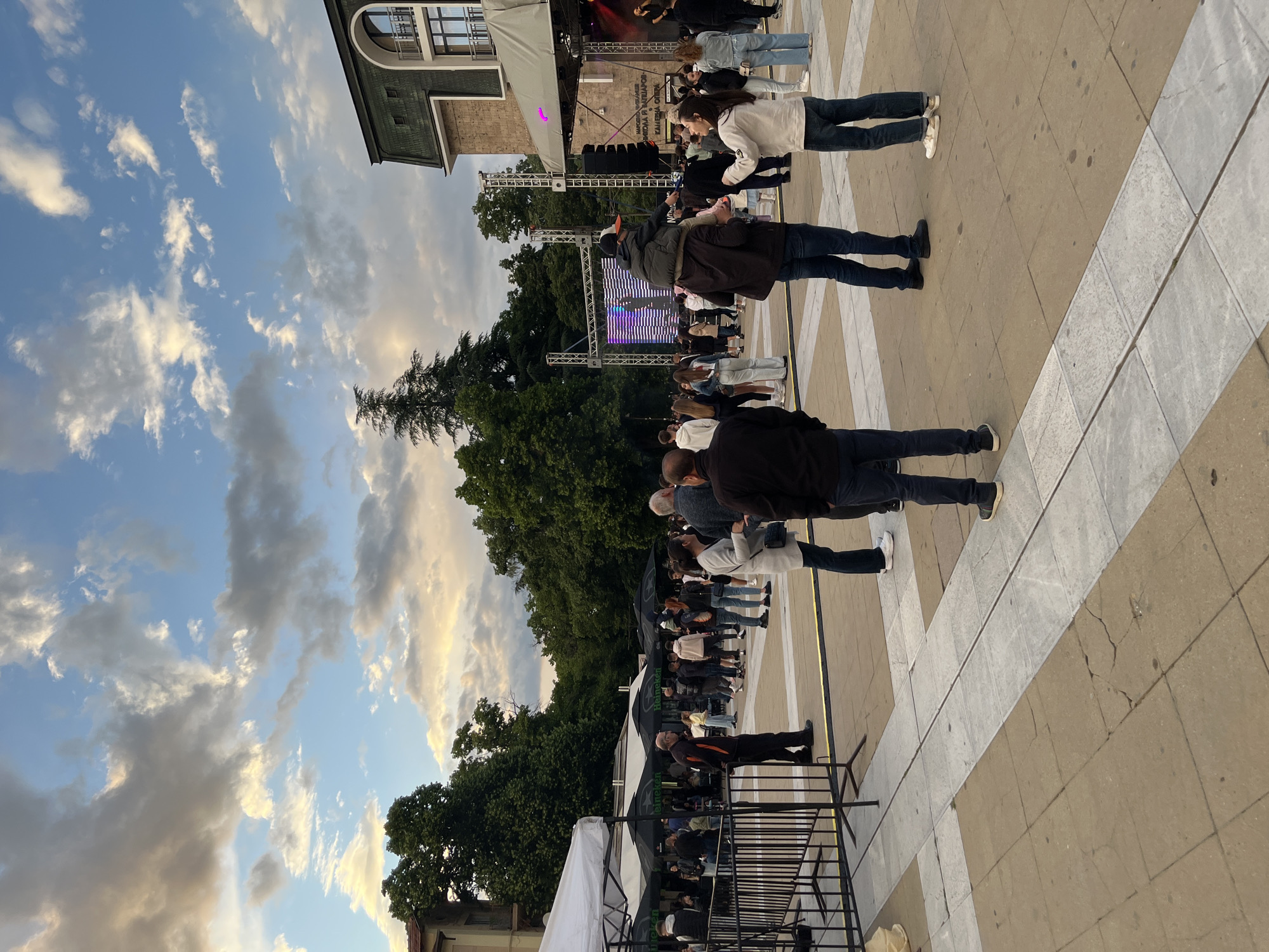 Here's a description of the image:
The image captures an outdoor scene featuring a crowd of people gathered in what appears to be a public square or plaza. In the background, a stage is set up, equipped with sound systems and lighting. The stage suggests a performance or event is taking place. The people in the foreground are facing the stage, and some are taking photos with their phones, which indicates that they are observing a performance. The surrounding environment includes trees, buildings, and a partly cloudy sky, suggesting it is either late in the afternoon or early evening. The ground is paved with geometric patterns. There is no indication of the location in the city of Unknown, Unknown, apart from what can be inferred from the building architecture and the vegetation.