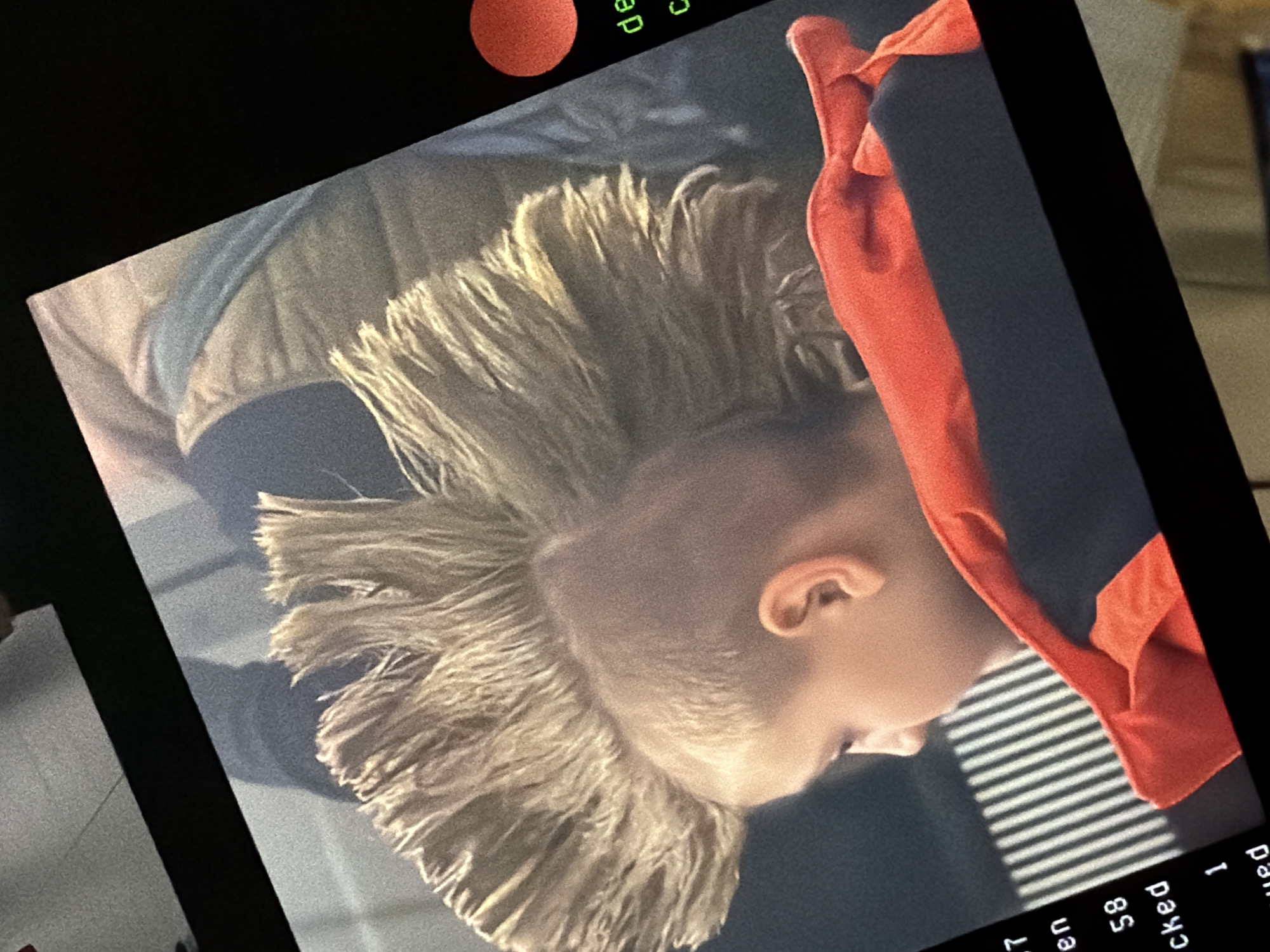 The image shows a close-up of a person's head with spiky, blonde hair. The person is wearing a red and black garment, possibly a hoodie or jacket. The background appears to show venetian blinds. Text is visible in the lower right corner, with words partially cut off such as "en", "cked", and "ed". A partially visible orange circle is present in the top left. There are no landmarks to suggest a location in Folkestone and Hythe District, United Kingdom. The image is likely a screen capture of a video.