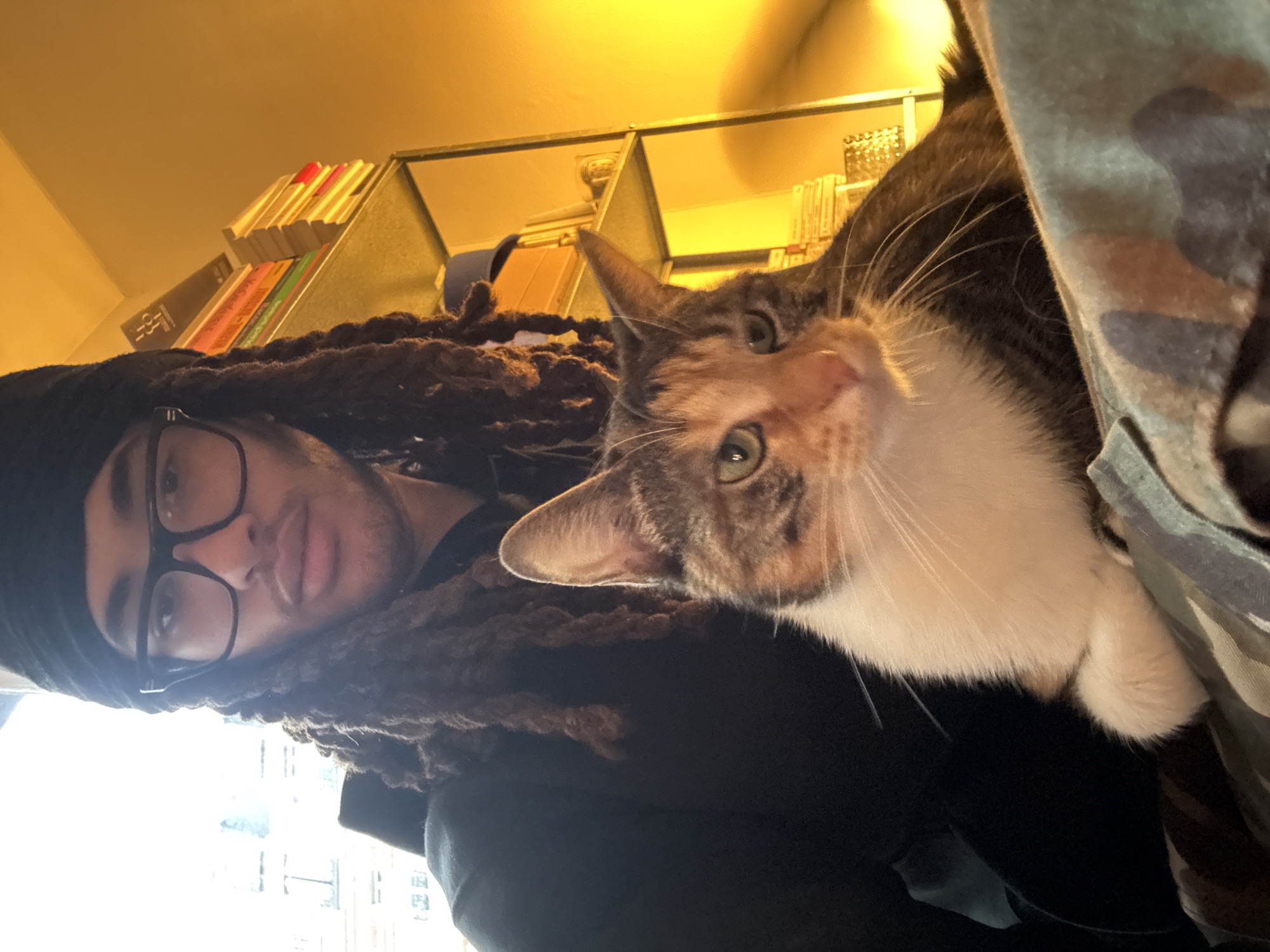 A selfie shot in a well-lit room, possibly during the daytime, features a man with dreadlocks and black-rimmed glasses on the left. Next to him, a cat with a white chest and orange and brown patterned fur looks at the camera. The background reveals a bookshelf filled with books, and hints of a cluttered environment with a metal shelf above the books. While the image doesn't provide explicit indicators of Paris, France, the style of the shelf and books suggest a potentially urban interior typical of European cities like Paris.
