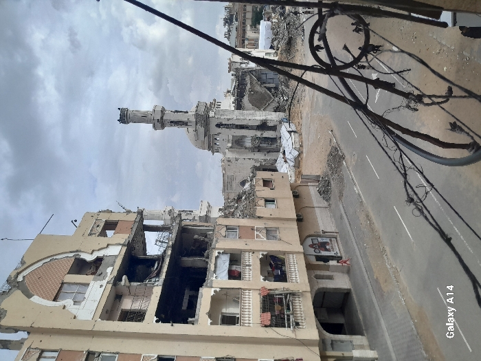 The image depicts a scene of extensive urban devastation in Khan Yunis, Palestinian Territory, under an overcast sky. In the immediate foreground, dark, tangled wires and a metal railing partially obstruct the view from an elevated vantage point. To the left, a multi-story building, appearing to be residential, exhibits severe structural damage, with several upper floors partially collapsed and interiors exposed; rebar is visible, and laundry hangs from a lower-level balcony. Debris piles are visible at the base of this building and along the adjacent sidewalk. A single small figure, dressed in red, walks rightward on the sidewalk. An asphalt road with faded white lane markings traverses the midground, bordered by dirt and rubble. To the right and in the background, a heavily damaged mosque structure features a prominent, partially intact minaret and a collapsed dome, surrounded by substantial rubble and the remnants of other destroyed buildings. Further in the background, more destroyed structures and several makeshift shelters constructed from tarps or corrugated metal are visible. A small group of approximately three to four indistinct figures walks on a distant dirt path on the far right.