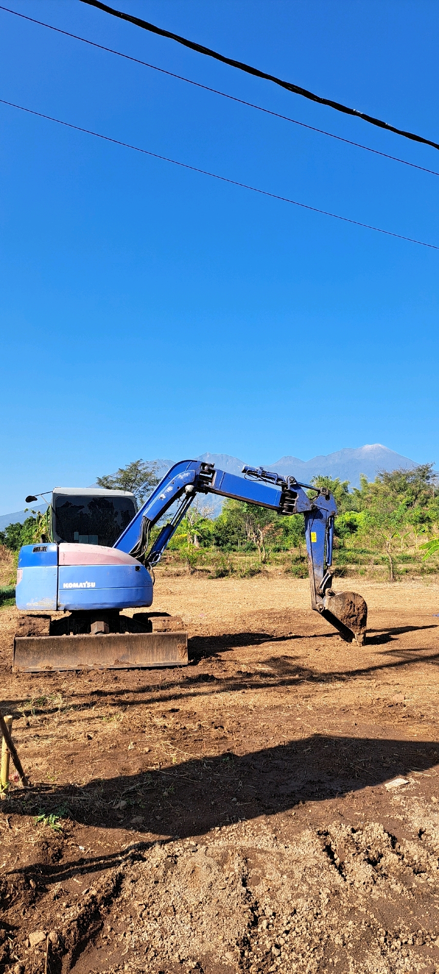 In an open field under a clear blue sky in Sumber Suko, Indonesia, a blue Komatsu excavator sits on a dirt surface. The excavator's arm is extended, with its bucket holding a clump of dirt. In the background, a range of green trees and mountains are visible, with a snow-capped peak in the distance. Power lines run across the top of the image. The ground around the excavator is dry and rough, with some scattered dry grass and evidence of recent digging. The scene appears to be a construction or land-clearing site.