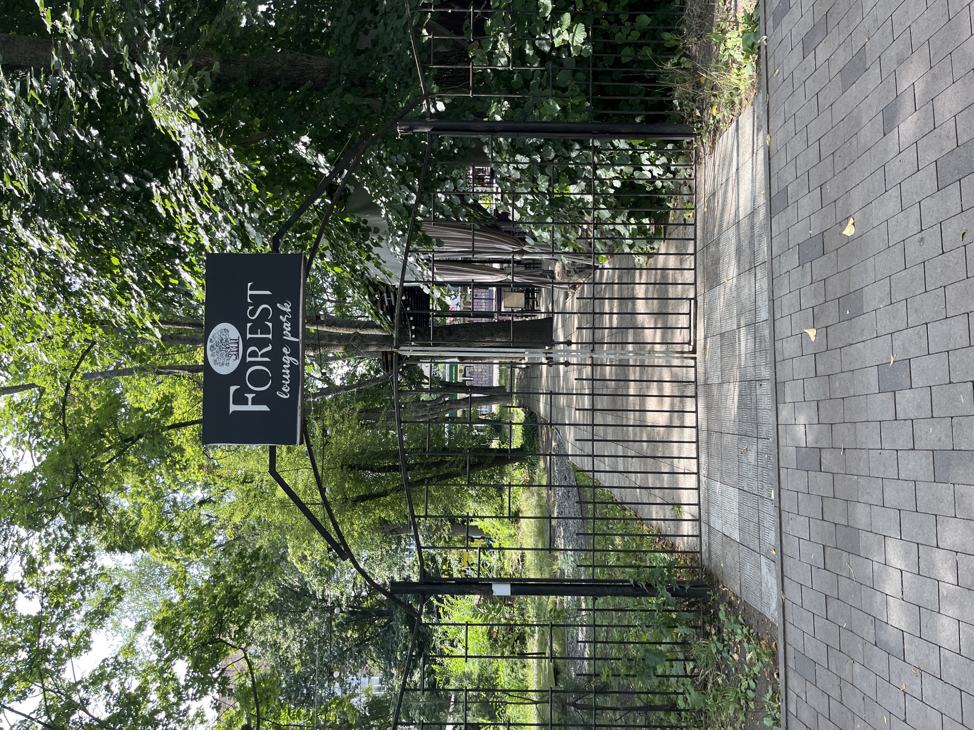 The image shows a black metal gate and fence in front of a path leading into a park-like area with trees and foliage. A black sign above the gate reads "FOREST lounge park". The path is paved with grey rectangular stones. Beyond the gate, the path continues into a shaded area with trees and some tables and chairs, suggesting an outdoor seating area for a cafe or restaurant. The environment appears to be an urban park or a restaurant with an outdoor seating area. No people are visible in the image. The scene is lit by natural daylight. The location is the entrance to the "Forest Lounge Park" cafe, located in Kharkiv, Ukraine.