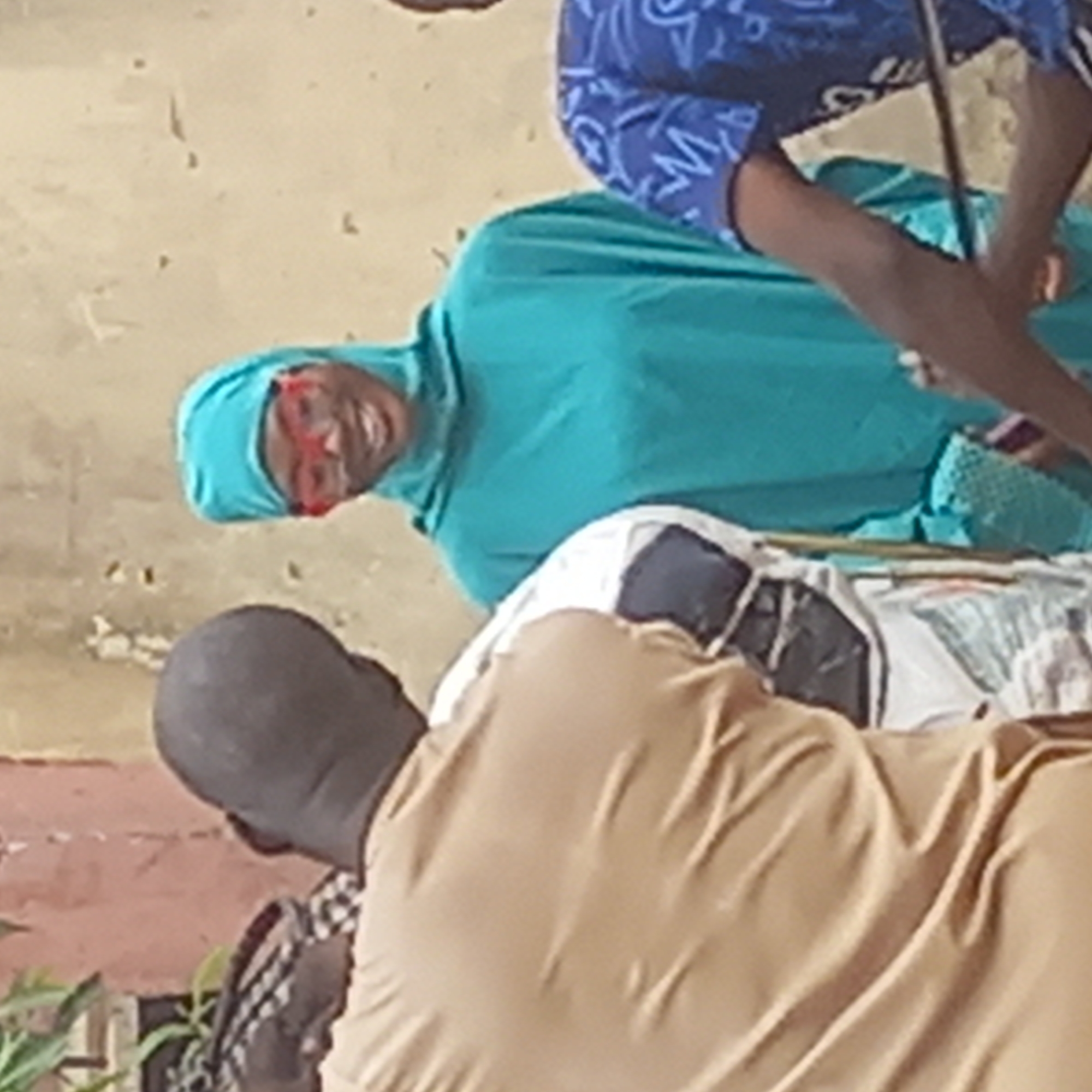 The image shows a woman in a teal hijab and red glasses looking up and smiling. A person wearing a blue patterned shirt stands behind her. A man with a shaved head and a tan-colored shirt is in the foreground. The location is Dadi Riba, Nigeria. The environment is somewhat cluttered, and a chair is partially visible.