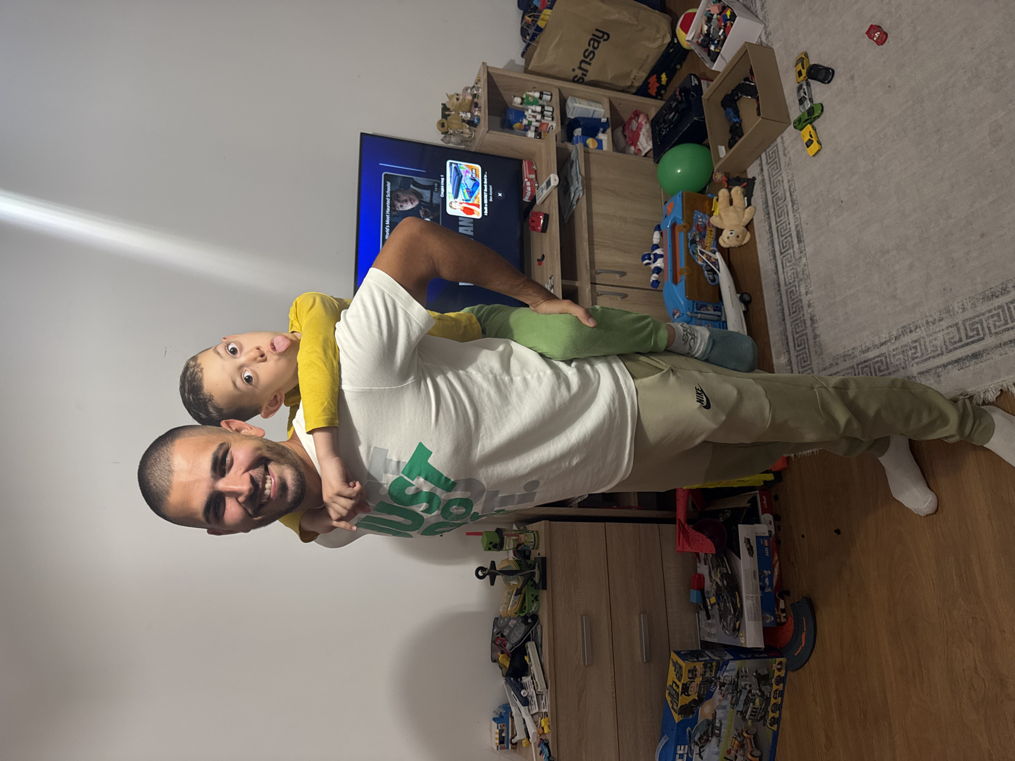 The image shows a man giving a piggyback ride to a young boy in a room filled with toys. The man is smiling and wearing a white t-shirt and light green pants. The boy is wearing a yellow shirt and sticking his tongue out. A television displays content on a shelf, which sits above the man and boy. Various toys are scattered around the room, including building blocks, cars, and a teddy bear. There are also some cabinets and shelves in the background. The image does not provide information about the location in Burgas, Bulgaria, nor can it be determined from the image contents.
