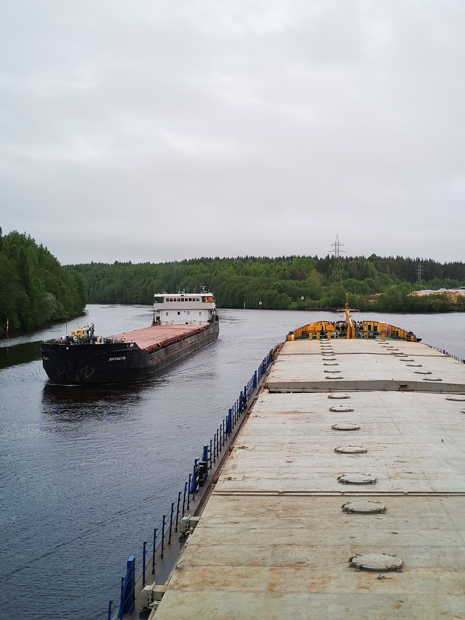 The image depicts a river scene in Depo, Russia, under an overcast sky. Two barges are visible. The first barge is black and carries a reddish-brown load. The name "Volgodonsk" is written in Cyrillic letters on its side. The second barge, positioned closer to the viewer, has a gray deck with a series of oval hatches and yellow equipment at its bow. Both barges are on a river bordered by green trees on either side. A power line is visible in the distance.