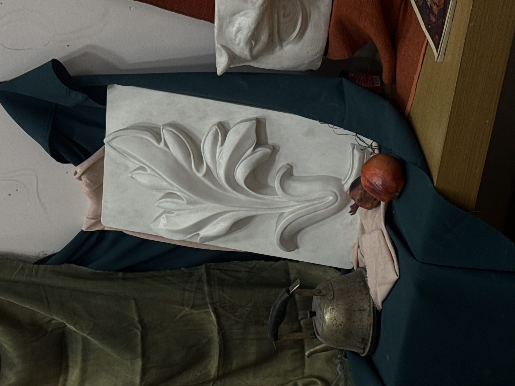 The image depicts a still life arrangement. Dominating the center is a rectangular plaster relief of an acanthus leaf design. To the upper left, a smaller plaster relief of a human face is visible. A dark teal fabric is draped around the reliefs. An orange-red pomegranate sits near the edge of the acanthus leaf relief. A tarnished metal kettle rests at the bottom of the frame, partially obscured by a green fabric. A peach-colored cloth is also visible. A wooden surface and edge of a framed picture are located to the right of the frame. The image's location is potentially Yerevan, Armenia.
