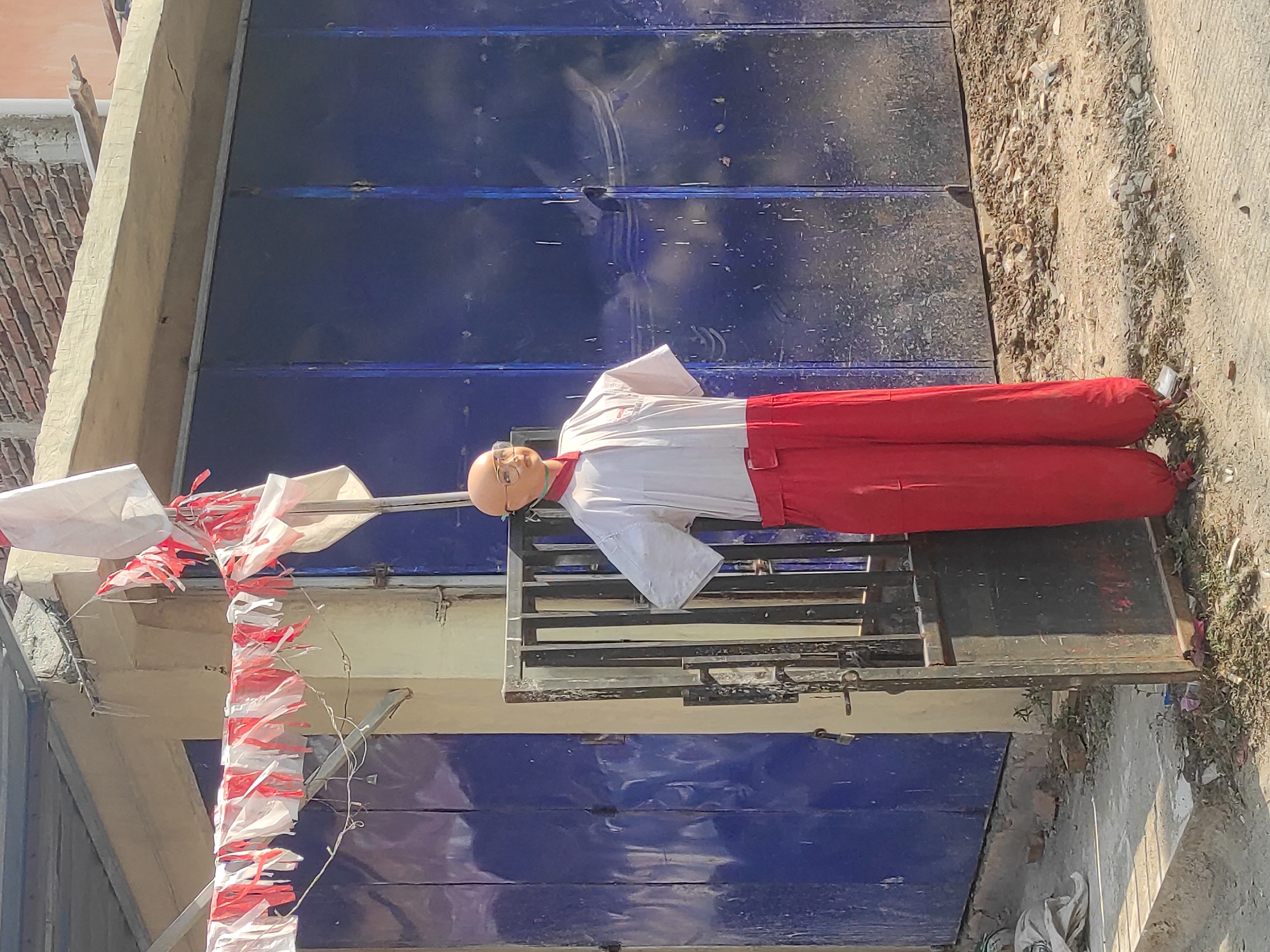A headless mannequin dressed in a white shirt and red pants stands in front of a blue corrugated metal door in Cankuang, Indonesia. The mannequin has a bald head with glasses and its hands are on its hips. Red and white streamers hang above it. The scene is outdoors with a brick building in the background. The ground is dirt and debris.
