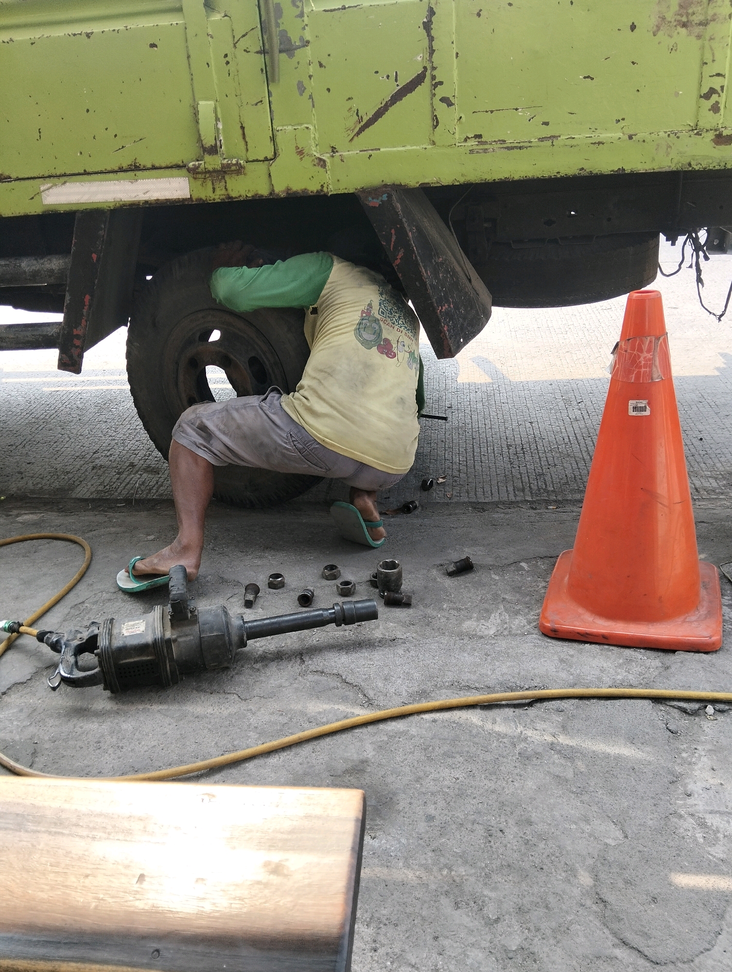 In the city of Semarang, Indonesia, a mechanic is seen working on the tire of a large, lime green truck. The mechanic, wearing a light yellow t-shirt and grey shorts, is crouched down beneath the truck, with his head and torso obscured by the vehicle. His legs are visible, clad in light blue flip-flops. To the right of the truck, an orange traffic cone stands on the ground, its surface showing signs of wear and tear, with some tape applied. A black air-powered impact wrench and several lug nuts are scattered on the cracked asphalt ground in the foreground, indicating the ongoing tire repair. A yellow air hose snakes across the ground, connecting to the impact wrench. The truck itself appears to be an older model, with visible rust and chipped paint, suggesting it has seen considerable use. The scene is illuminated by natural daylight, casting shadows on the ground.