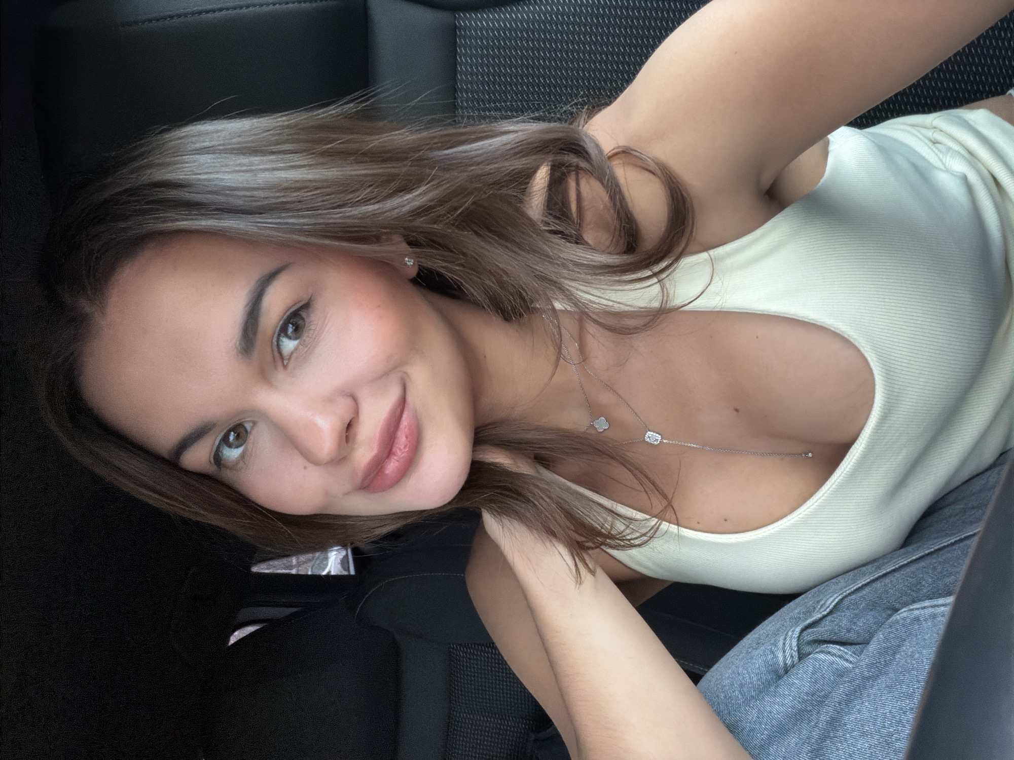 A woman is in the driver's seat of a car. She is fair-skinned, with brown hair, light brown eyes, and pink lipstick. She is wearing a white tank top and blue jeans. A silver necklace is visible. The car's interior is dark. The photo was not taken in Krasnoyarsk, Russia.