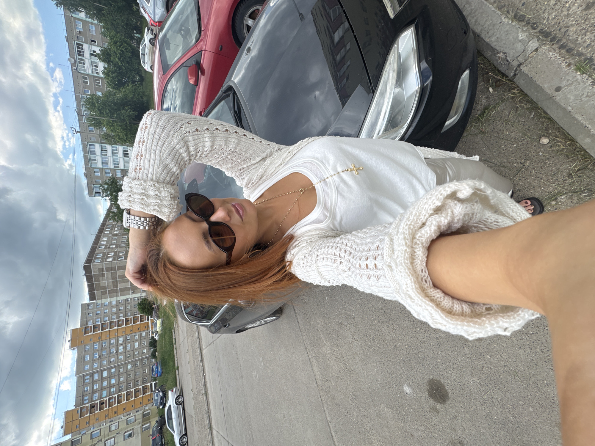An outdoor, eye-level, medium shot captures a woman with auburn hair taking a selfie in front of a black car. She is wearing oversized sunglasses, a white tank top, a white crocheted cardigan with bell sleeves, and light-colored pants. A gold cross necklace with a long chain is visible. Her right arm is raised, with her hand touching her hair. A large, shiny, black car is parked next to her, with its front bumper and headlight visible. A red car is parked behind the black car. In the background, several residential buildings are visible, characteristic of Chaikovsky, Russia. The buildings are multi-story, with a mix of apartments and balconies. The sky is partly cloudy, with gray clouds dominating the upper portion of the frame. The ground is paved with concrete and asphalt, with some grass and debris visible near the curb.