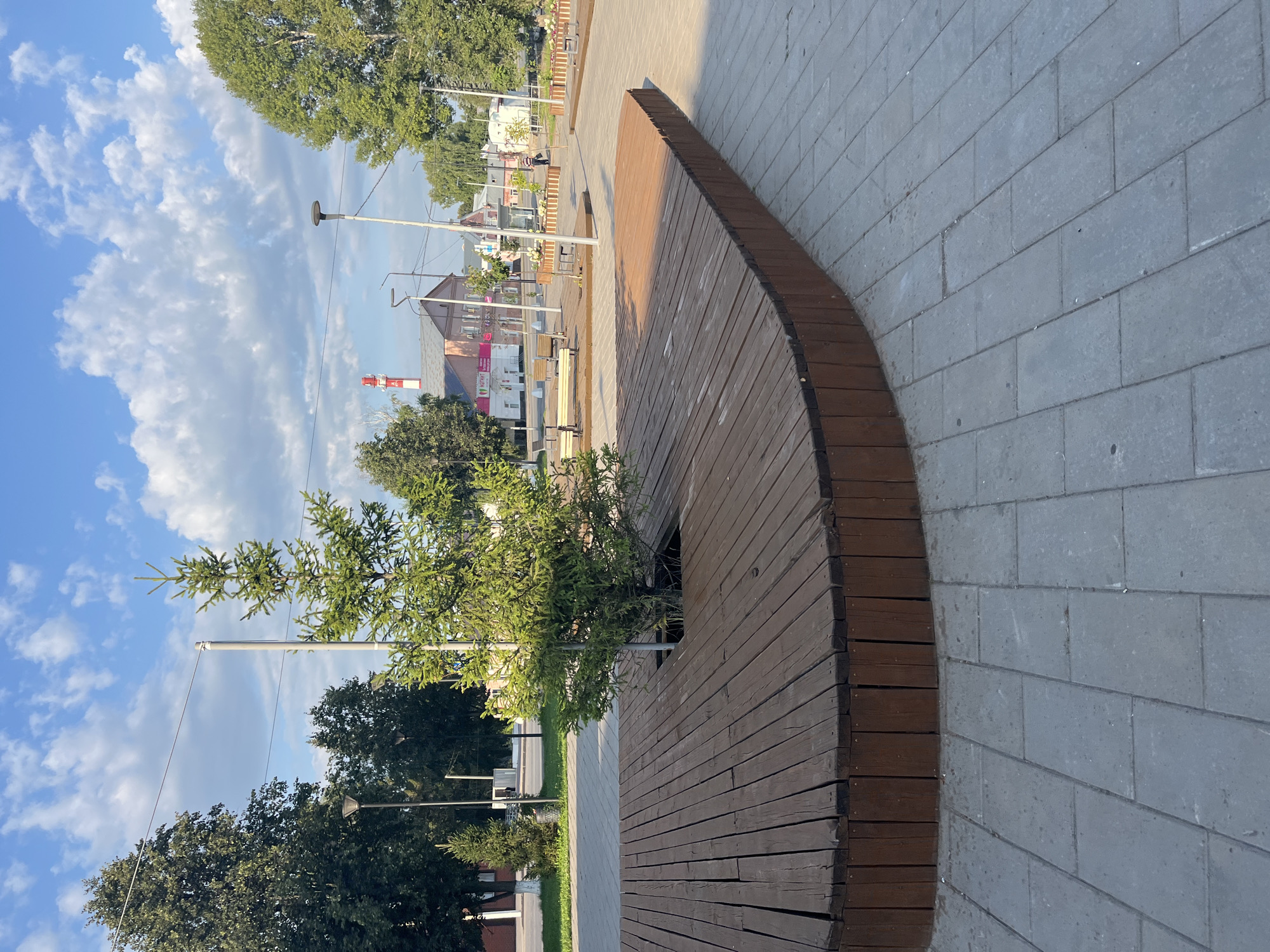 A medium shot captures a public outdoor space with a large, curved wooden bench in the foreground. The bench is constructed from parallel wooden planks, with a darker brown wooden base. A small evergreen tree is planted in a circular opening in the center of the bench. The ground is paved with grey rectangular tiles. In the background, there are more trees, streetlights, and buildings, including a pink building with a red and white striped chimney. The sky is blue with scattered white clouds. A person is visible walking in the distance. The scene appears to be in Novye Turbasly, Russia, a location suggested by the context.
