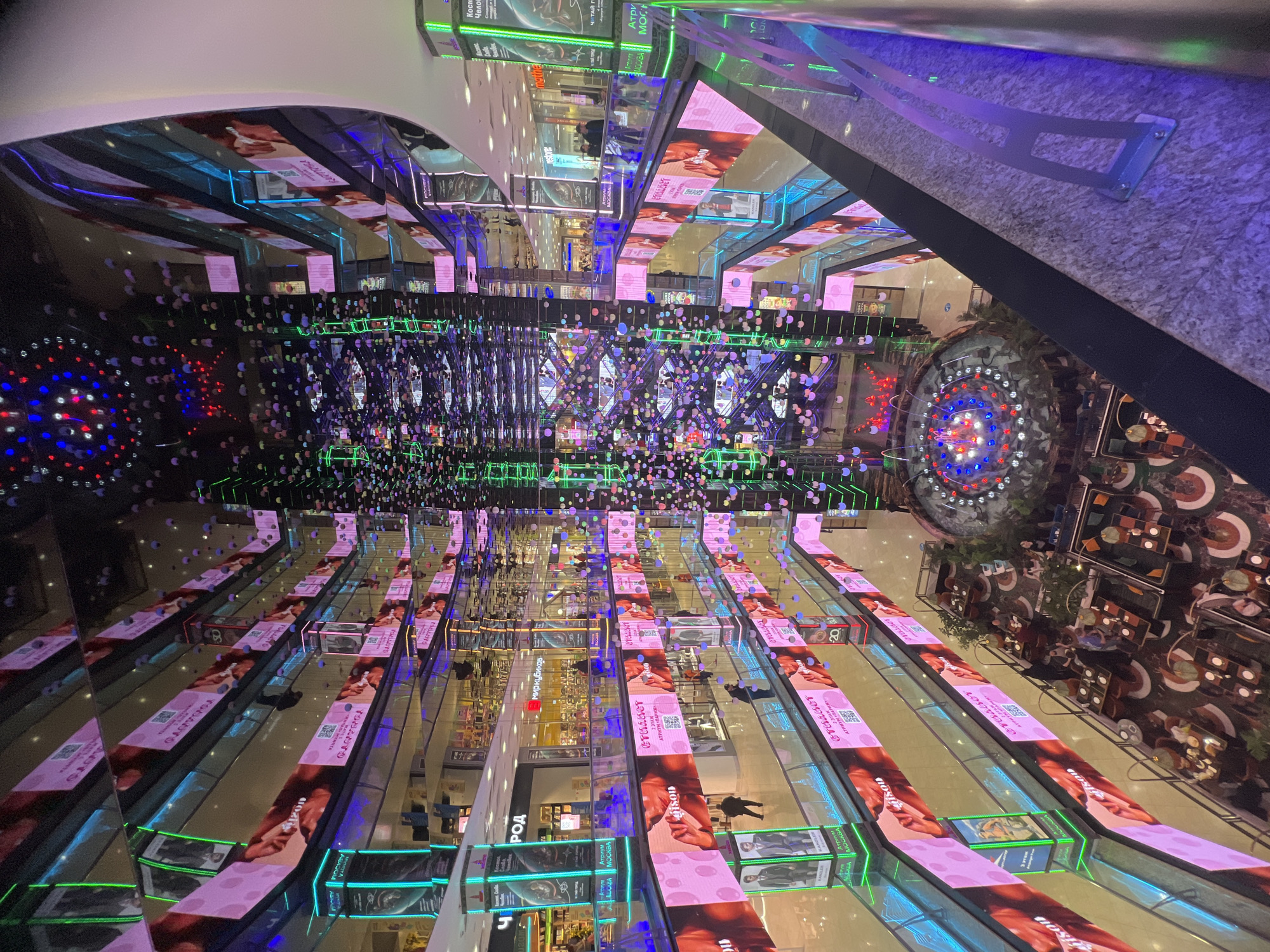 Captured from an elevated perspective, the photograph showcases the multi-story expanse of a shopping mall located in Moscow, Russia. The mall is adorned with digital displays exhibiting a variety of advertisements, interspersed with retail signage visible on the store fronts. Several individuals are observed moving throughout the mall's walkways. A central display, featuring illuminated orbs in red, white, and blue, serves as a focal point. There is a cafe located at the edge of the shopping mall. The mall's architecture includes glass railings.