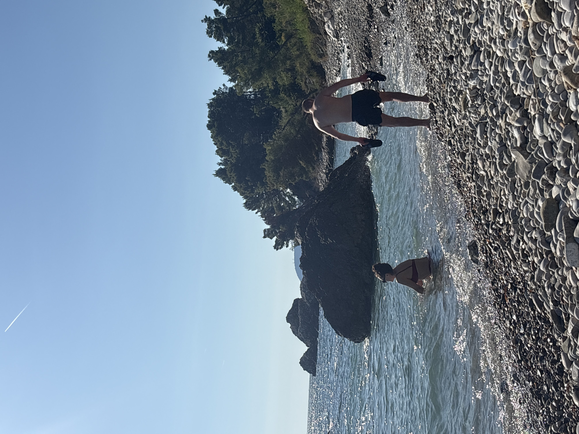 The image shows a rocky beach on a sunny day. A man in black shorts stands at the edge of the water, holding sandals in his hands, near a rock formation. A woman is standing in the water further down the shoreline. Behind them is a line of trees and a clear blue sky with a contrail visible. The location is likely near or in the city of Bar, Montenegro, given the rocky coastline, vegetation, and clear weather typical of the region. The beach is covered in smooth, rounded stones. The water is clear with some visible wave action.