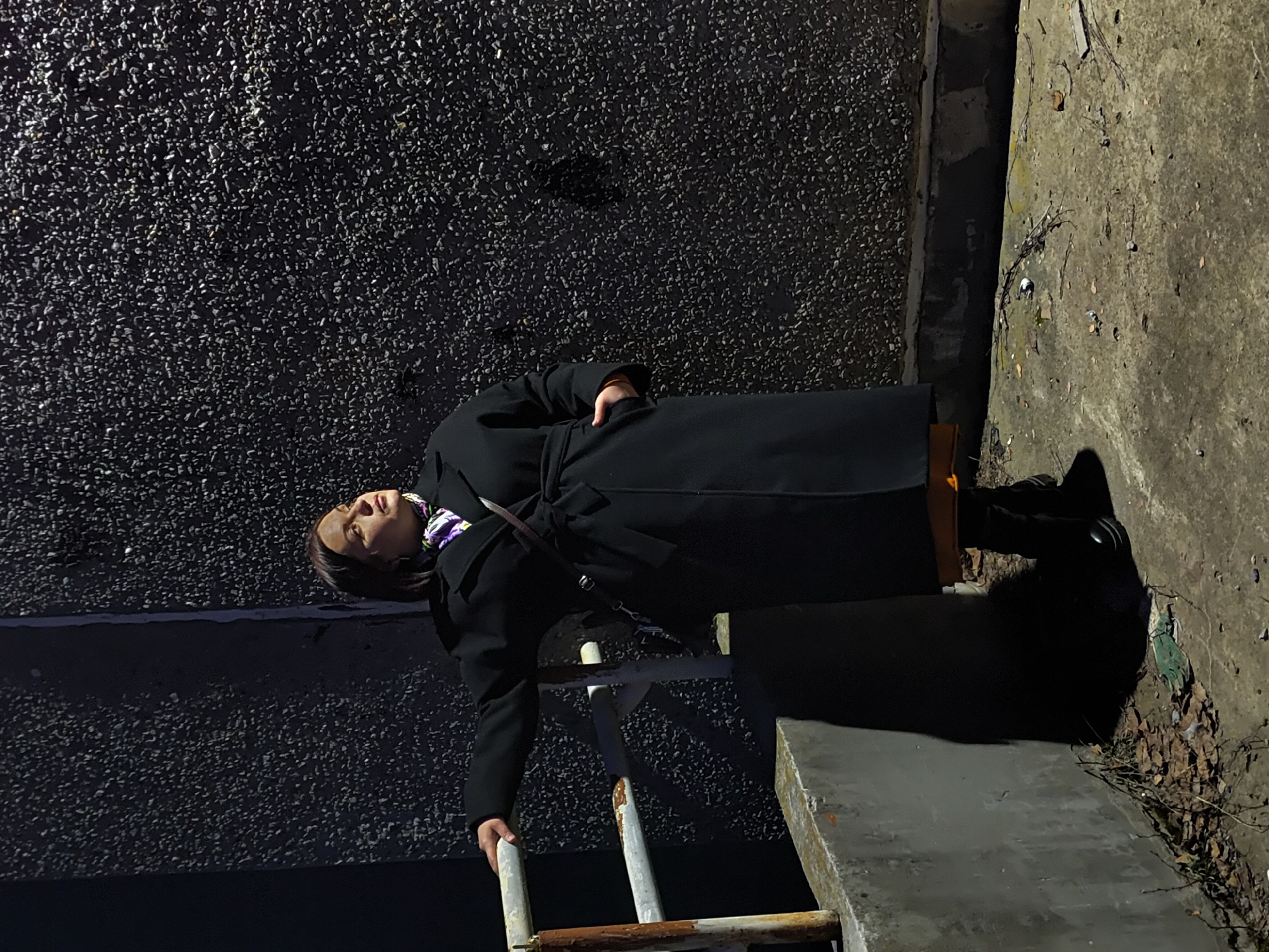 A person wearing a black coat and shoes is standing on a rusty, white ladder in an urban environment. The person's left hand is holding the ladder while their right hand is inside their coat pocket. The ladder is positioned against a dark, textured wall and a concrete ledge. The environment includes dry leaves and debris. The city is Марковский, Russia.