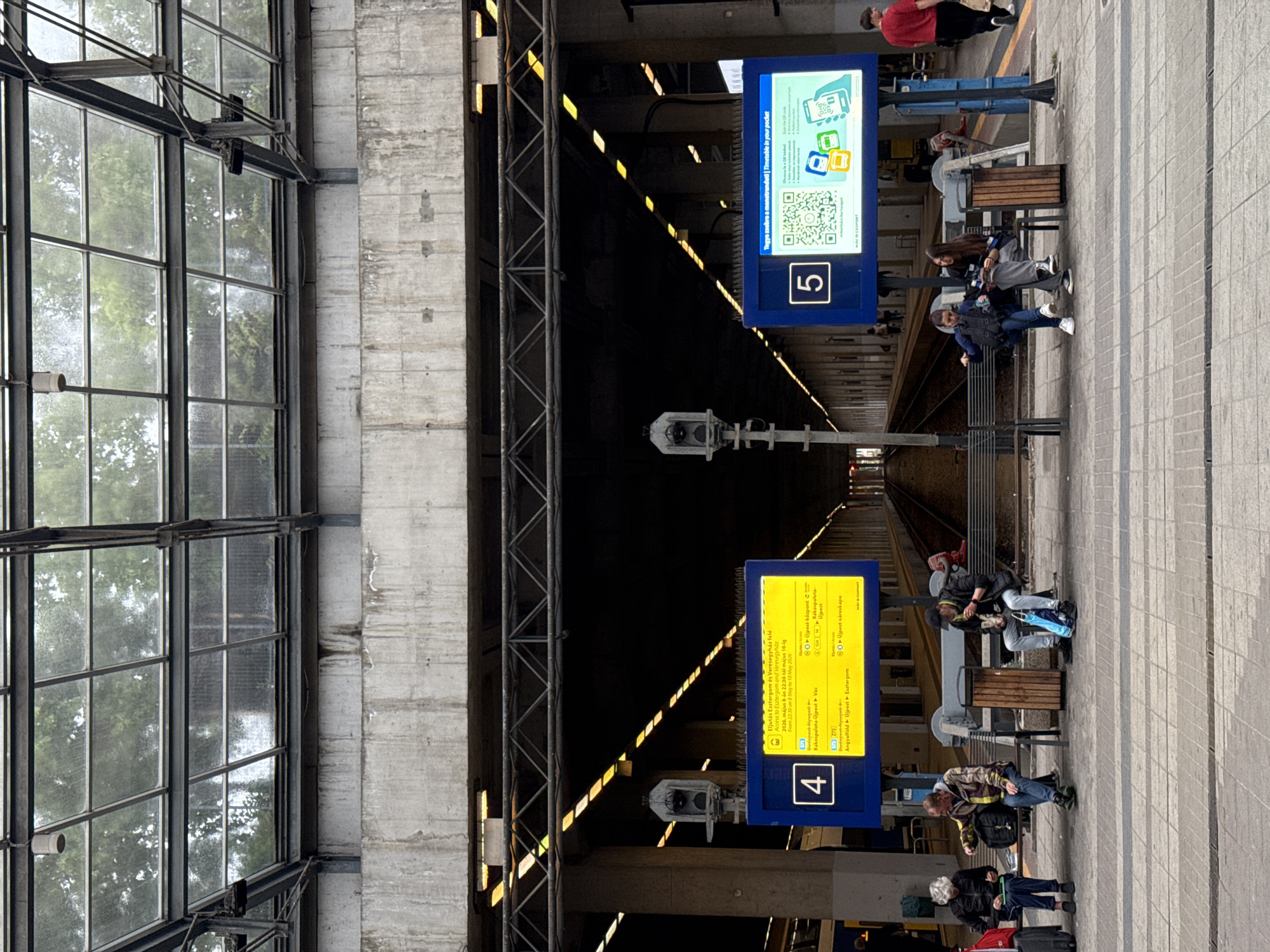 The image captures a scene at what appears to be the Budapest-Nyugati railway station in Budapest, Hungary. Two digital platform displays are visible: one indicating platform 4, with information in Hungarian about trains to Esztergom and Veresegyház. The other shows platform 5, offering timetable and QR code. Several people are waiting near platform 5, some seated on benches. The station's architecture includes a large glass roof and concrete structures. Railway tracks stretch into the distance, disappearing into a darkened area.