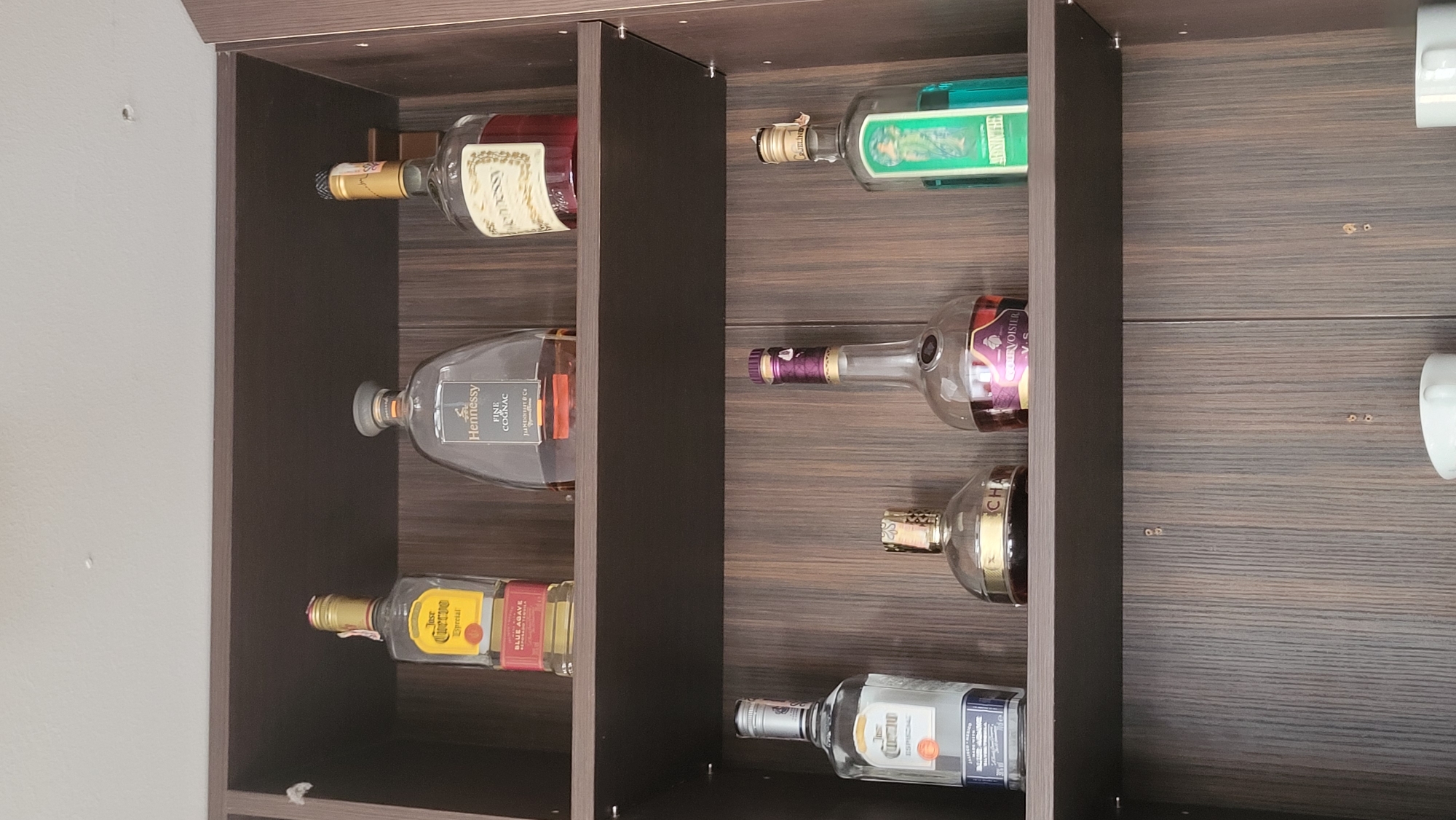The image shows a dark wood cabinet with three shelves. The top shelf holds three bottles of liquor: a bottle of Jose Cuervo Especial tequila, a large bottle of Hennessy Fine Cognac, and a bottle of Hennessy cognac with a red label. The middle shelf displays four bottles: a bottle of Jose Cuervo Especial, a bottle with a gold label, a bottle of Courvoisier V.S., and a green bottle with a label featuring a woman. The bottom of the cabinet shows a portion of two white mugs. The image does not contain any information about the city of Prešov, Slovakia.