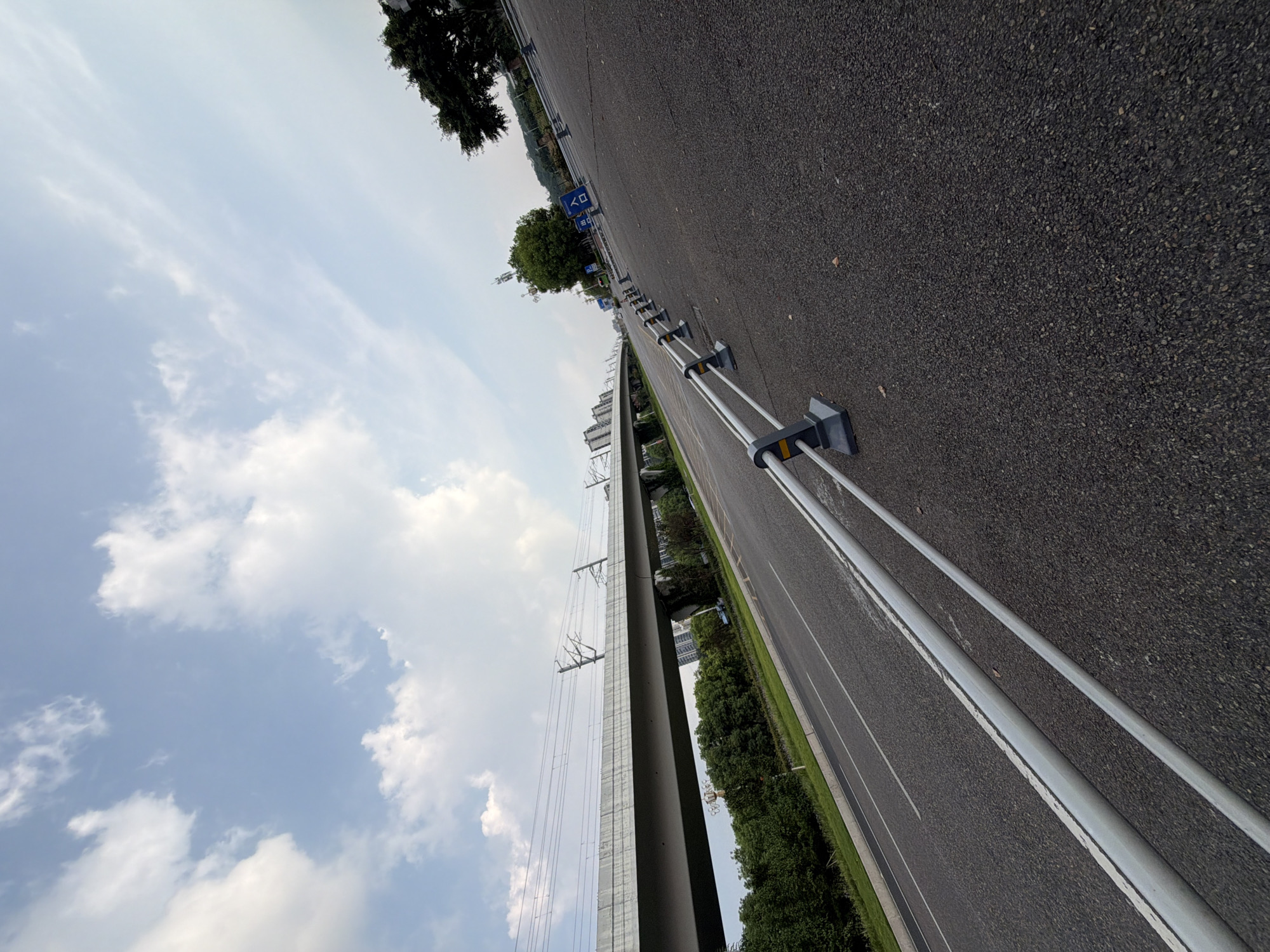 The image depicts an elevated railway or highway structure spanning across a road in Jiangjin, China. The foreground is dominated by a grey asphalt road with a white line marking. A metal guardrail with grey supports runs along the right side of the road, extending into the distance. In the midground, lush green trees line the left side of the road, with the elevated structure above them. The structure itself is a grey concrete overpass with visible overhead electrical lines, suggesting it's a railway or an electric highway. In the background, more buildings and trees are visible under a sky filled with scattered white clouds. The overall scene is one of urban infrastructure integrated with a natural landscape.