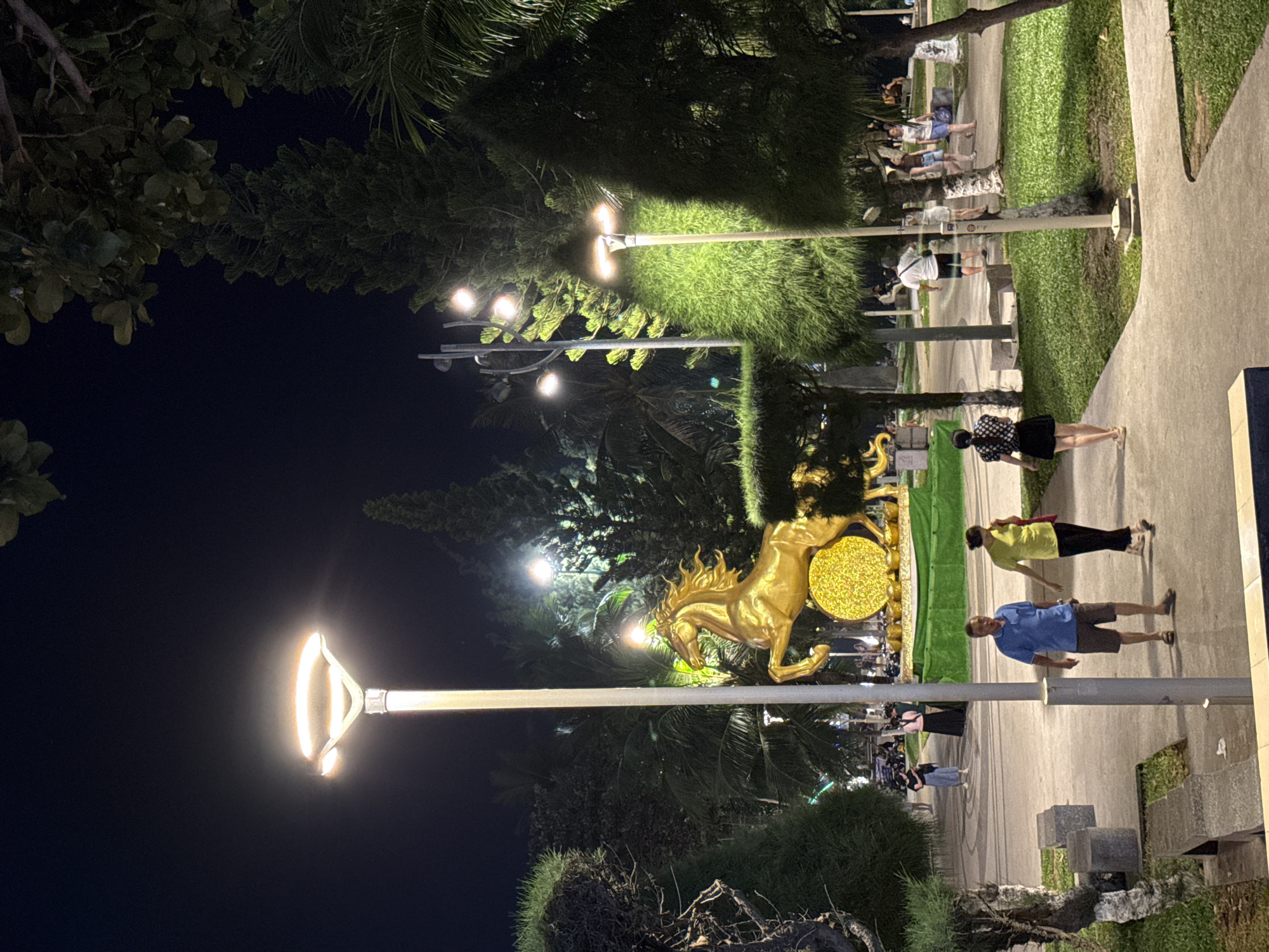 The image captures a nighttime scene in a park in Nha Trang, Vietnam, likely in a tourist area given the prominent golden horse statue. A tall, modern lamppost with a circular light fixture stands prominently on the left, casting a warm glow on the paved walkway and surrounding greenery. To the right of the lamppost is a large, ornate golden statue of a horse rearing up, with a circular, textured gold disc behind it, supported by a green base.
Several people are visible on the walkway. In the foreground, a man in a blue polo shirt and khaki shorts walks to the left, while a woman in a yellow t-shirt and black pants walks beside him to the right. Further back and to the left, a woman in a black top and a long black skirt stands facing away from the camera. Other individuals and small groups are scattered throughout the park, some walking, others appearing to be gathered or seated.
The park features well-maintained green spaces with manicured bushes and trees, including tall, slender pines and palm-like trees. The ground is a mix of paved paths and grassy areas. The overall ambiance is that of a public gathering space at night, illuminated by artificial lighting and creating a somewhat festive or celebratory atmosphere, possibly due to the presence of the striking golden statue. The dark sky suggests it is late evening or night.