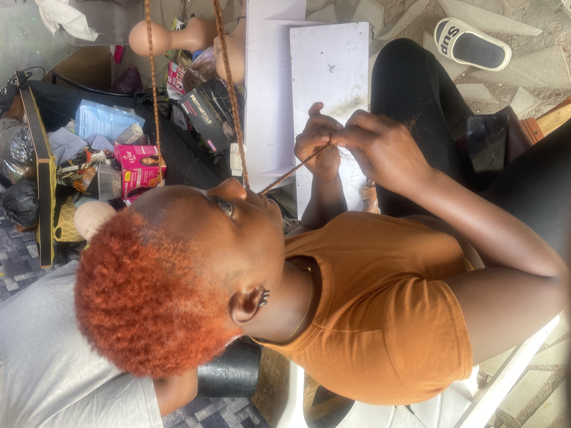 A high-angle shot shows a person with reddish-orange hair, wearing a mustard-colored shirt and seated in a white plastic chair, braiding hair extensions. The person is dark-skinned and has an earring in their left ear. Their right hand is braiding the extension, while the left hand holds the braid in place. A white sheet of paper lies on the chair. The background includes salon supplies, such as wigs, mannequin heads, hair products, and a storage cabinet with various objects. A white sandal with black "SUP" letters is on the floor. The ground appears to be a tiled patio. Location is inferred to be in Jos, Nigeria.