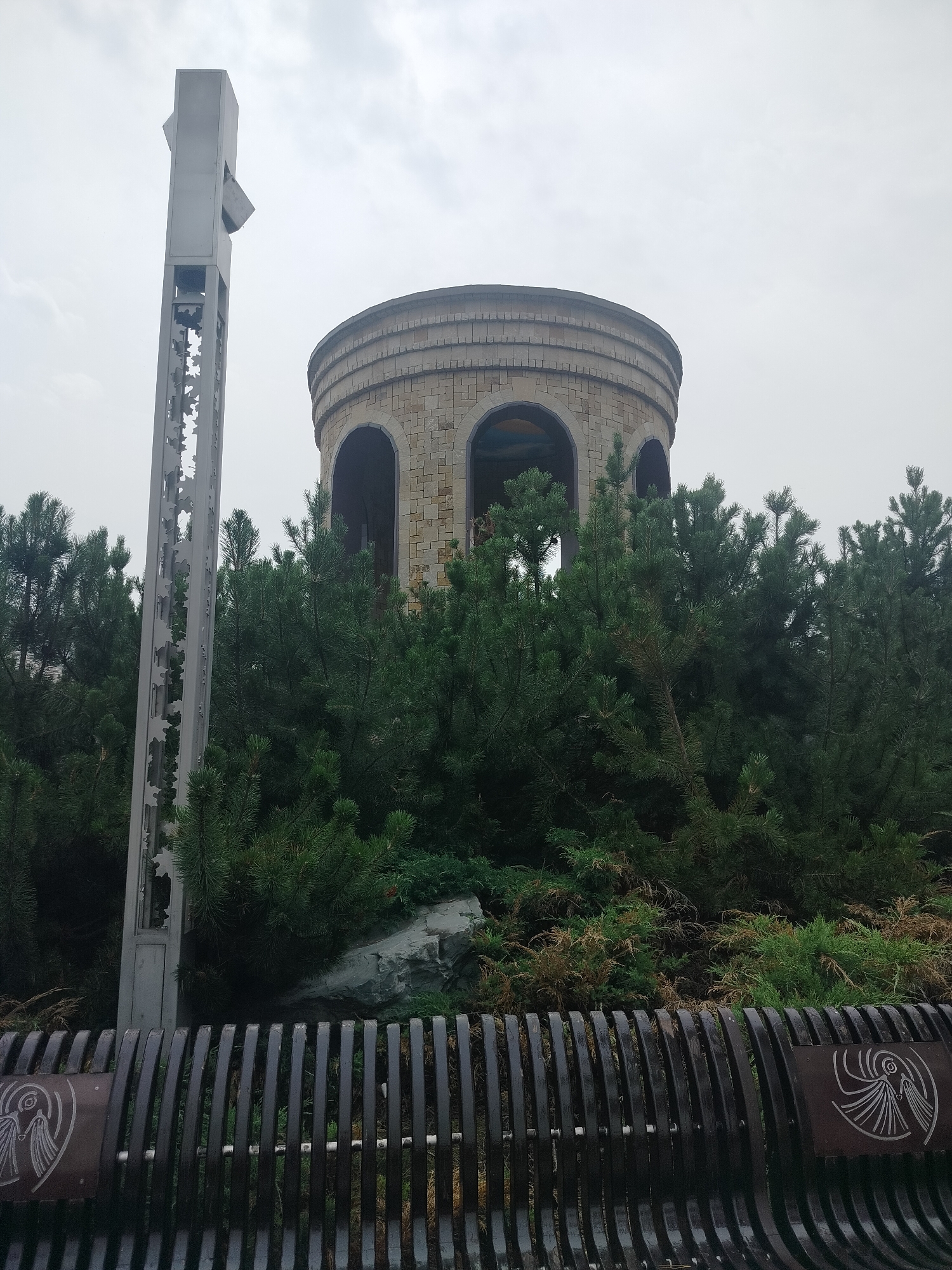 The image depicts a cylindrical stone building with arched openings, possibly a historical monument or observation tower, nestled amidst dense evergreen trees. To the left of the structure stands a tall, angular metallic sculpture or monument, featuring an intricate cut-out design along its length. In the foreground, a dark, slatted bench with decorative metallic plaques is visible. The sky is overcast and hazy. This scene is located at the Monument to the Victims of Political Repressions in Kemerovo, Russia, a site dedicated to commemorating individuals who suffered under political repression.