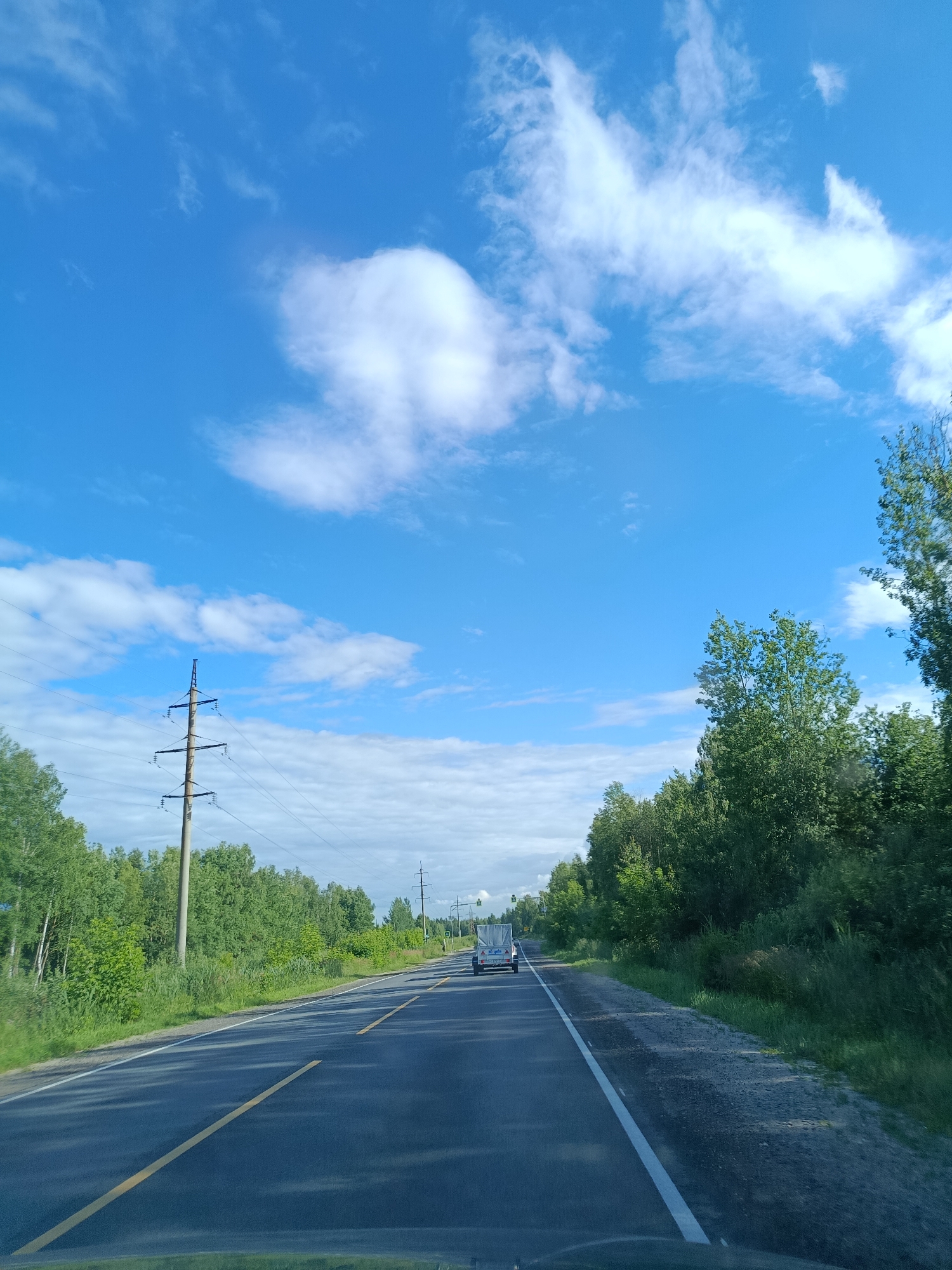 This media file depicts a scenic view from a vehicle driving on a paved road in Тополиный, Russia. The sky is a vibrant blue, dotted with fluffy white clouds, suggesting fair weather. The sun appears to be high in the sky, indicating it is likely daytime, possibly late morning or afternoon.
The road, lined with a solid yellow line on the left and a white line on the right, curves gently into the distance. On either side of the road, lush green trees and foliage form a dense forest, with some taller trees on the right side of the frame. Electrical power lines and tall utility poles are visible along the left side of the road, a common sight in rural or semi-rural areas.
In the distance, a white trailer being towed by a vehicle is visible on the road, indicating that travel is taking place. The overall atmosphere is serene and tranquil, evoking a sense of a peaceful journey through the Russian countryside. No people are clearly discernible in the image. There is no visible text within the frame.
