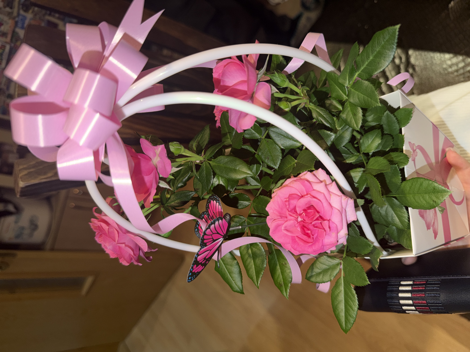 A close-up shot features a bouquet of pink roses, arranged within a white, curved frame. The arrangement is adorned with a large pink bow and a decorative butterfly. The roses, in various stages of bloom, are interspersed with green leaves. The bouquet is held in a white gift box with a pink bow design. The background shows a wood-toned surface with other items, and the edge of a person's hand can be seen holding the gift box. The image was possibly taken indoors under artificial lighting. Location details (city of Buhovo, Bulgaria) are not evident in the image.
