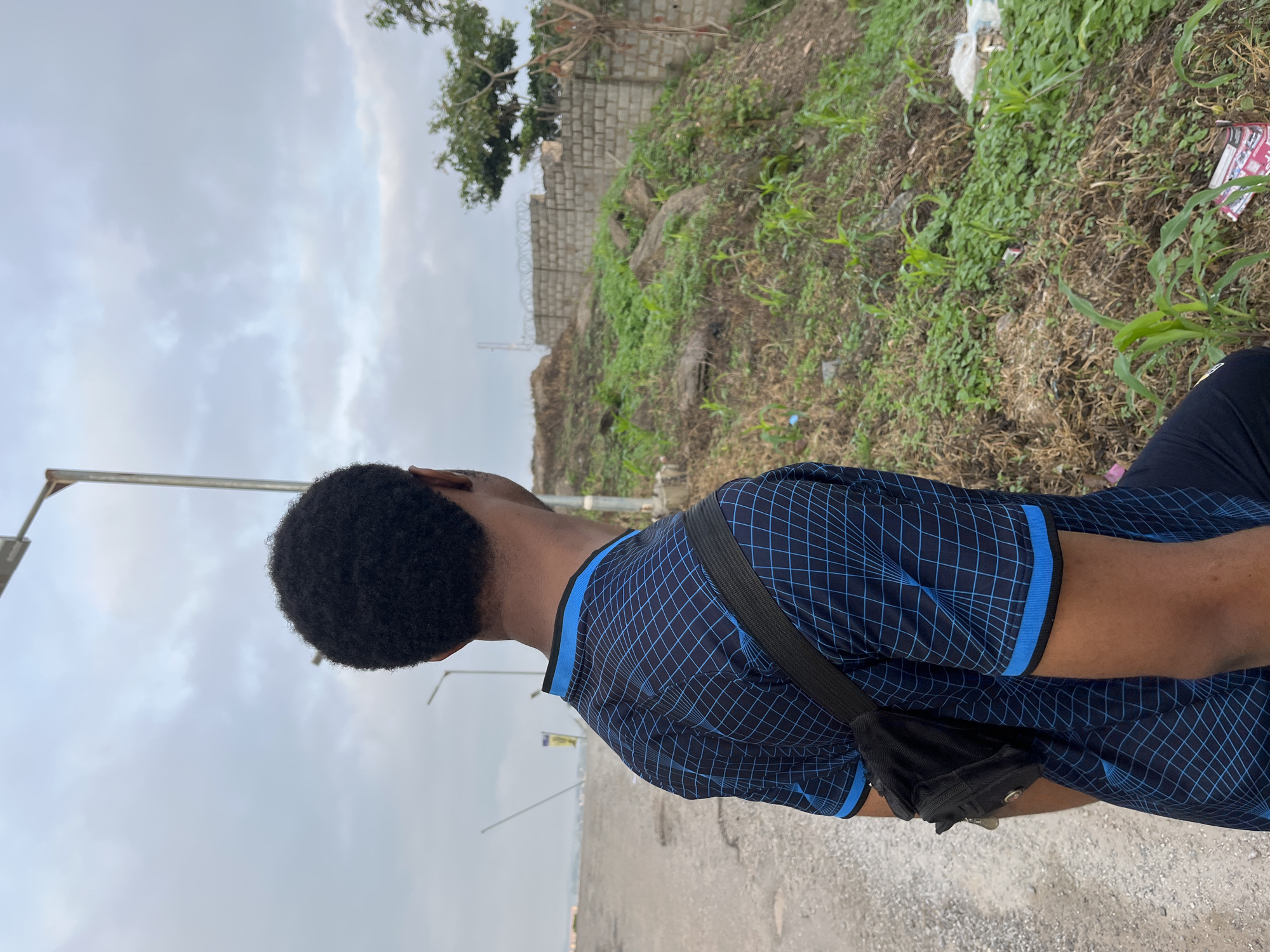 An over-the-shoulder, medium shot captures a person from the back, facing away from the camera. The individual is wearing a blue t-shirt with a grid pattern and a black fanny pack slung across their chest. They stand on a dirt and gravel road, with an overgrown, rocky embankment to their right. The embankment is covered in various green plants and some dried grasses. A stone wall with barbed wire fencing runs along the top of the embankment. In the background, the sky is overcast with gray clouds. A tall lamp post is visible to the left, and what appears to be a fence with a yellow sign can be seen further down the road. The overall setting suggests an outdoor, possibly undeveloped area within Abuja, Nigeria.