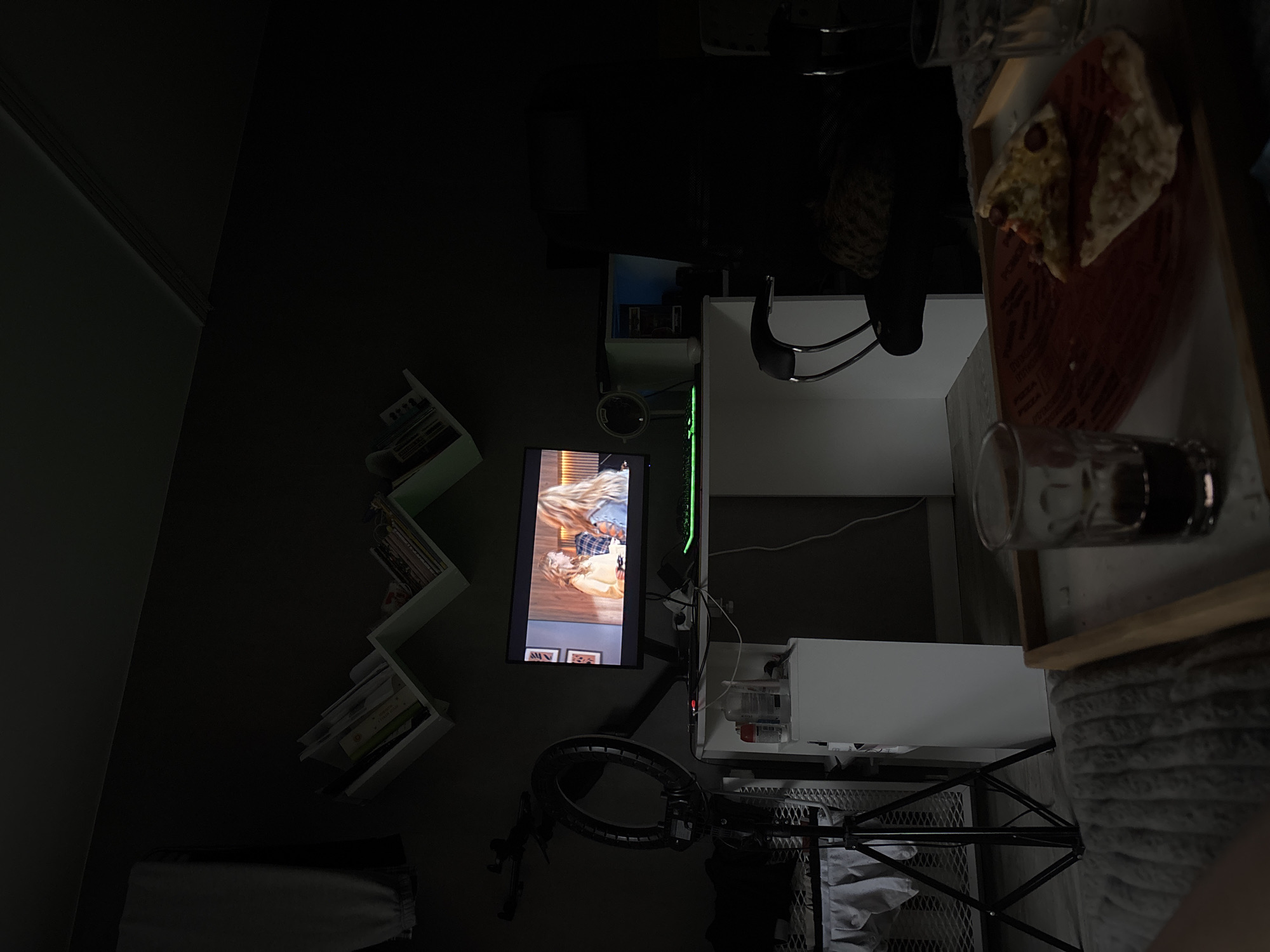 The image depicts a dimly lit bedroom interior. In the foreground, a wooden tray holds two slices of pizza and two glasses, one with a dark liquid and the other empty. A thick, grey blanket is visible under the tray. To the left, a ring light on a tripod stands beside a white wicker laundry basket. A white desk with a computer monitor, keyboard, and various items is centrally located. The monitor displays two women. The room is furnished with shelves on the wall holding books and decorative items. The overall ambiance suggests a personal living space, likely a bedroom or a room set up for content creation. The location of Chaikovsky, Russia is not deducible from the provided image.