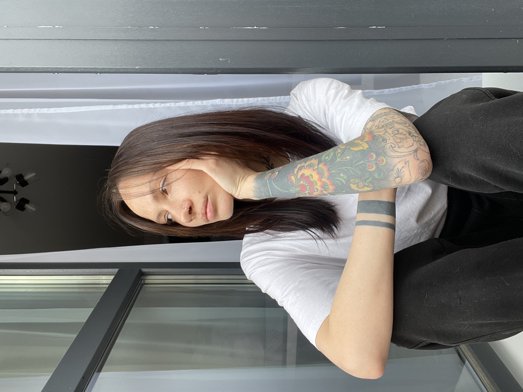 A young woman with dark hair and multiple tattoos is sitting, possibly on a windowsill, indoors. She is wearing a white t-shirt and black pants. Her left arm, adorned with colorful floral tattoos and two simple bands around her wrist, supports her head. She has a piercing in her nose. The background consists of a window with white curtains and an out-of-focus room interior, featuring a unique ceiling lamp. The location is possibly in the city of Pervomaysky, Russia, based on the request. The scene appears introspective and natural.