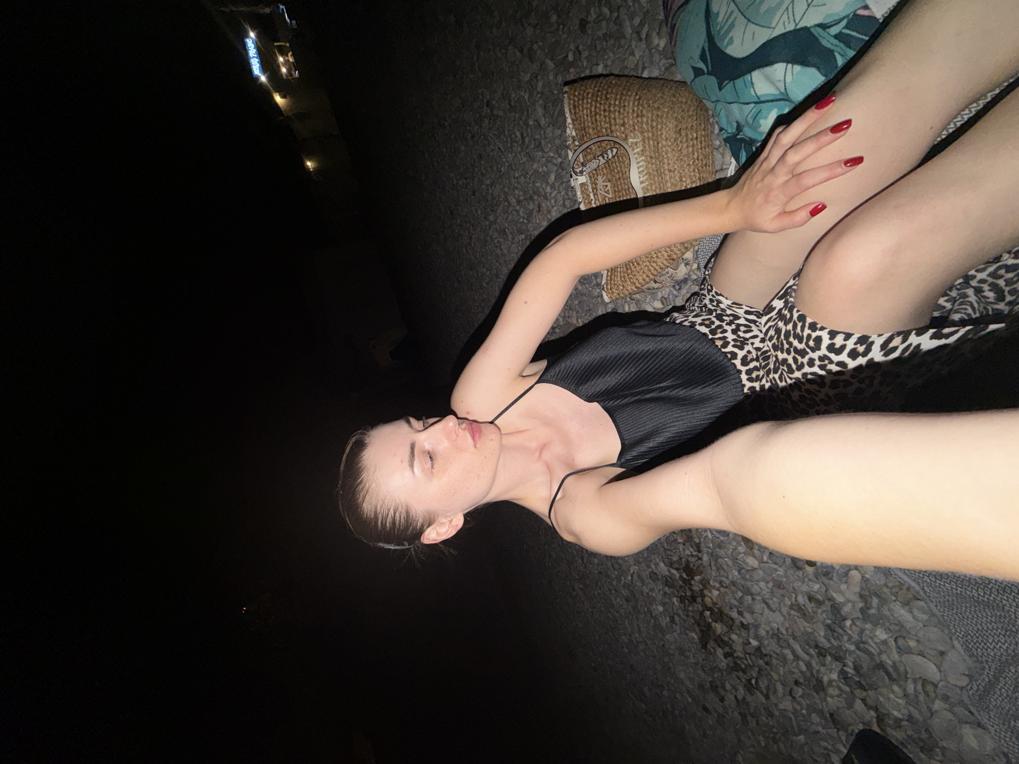 A woman is pictured at night, sitting on a pebble-covered surface, possibly a beach or riverbed. She is wearing a black camisole and a leopard-print skirt. Her hair is pulled back in a bun. A woven straw bag and a patterned blanket are near her. In the background, distant lights and what appears to be a sign are visible, suggesting an urban or resort area.