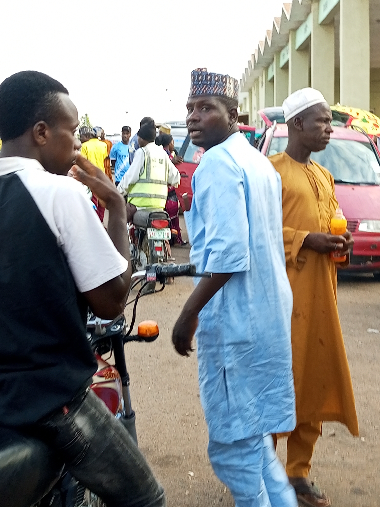 The image depicts a street scene in Gombe, Nigeria, likely near a market or transport hub given the presence of vehicles and pedestrians. In the foreground, a person wearing a black and white shirt is seated on a motorcycle, with their hands on the handlebars. To their right, another person in a light blue traditional garment and patterned cap is walking, facing away from the camera. Further to the right, a man in a mustard yellow garment and white cap is holding an orange drink bottle. In the background, a red car and several other individuals are visible. The environment appears to be a dusty, unpaved road in front of a building with a distinctive sawtooth roofline.