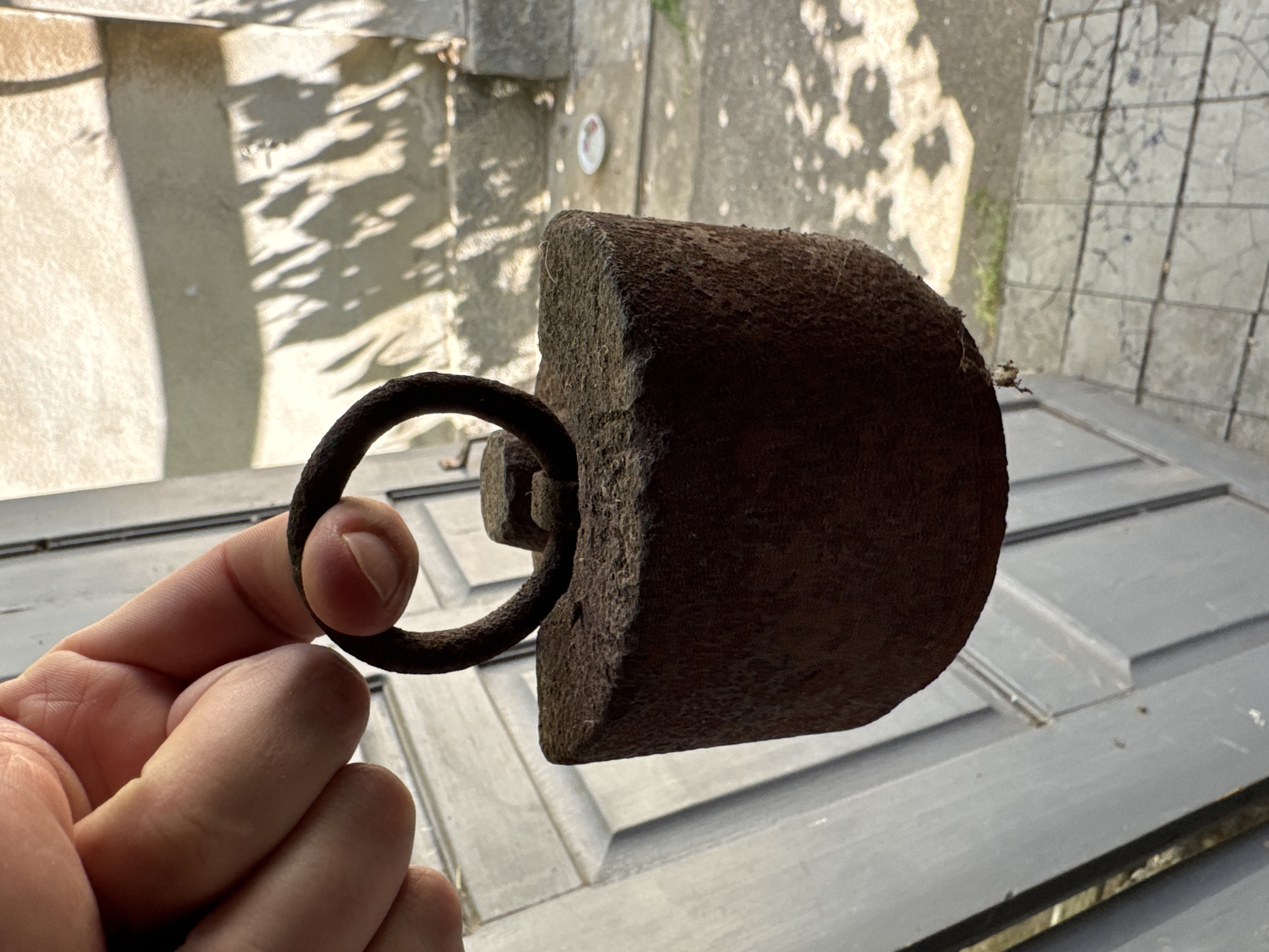The image shows a close-up of a hand holding a rusted metal object, likely a weight or a part of a tool. The object is dark brown and appears to be made of cast iron, with a rough, pitted texture. It has a D-shaped ring attached to one end, through which a finger is inserted. The background is somewhat blurred but appears to be an outdoor setting with dappled sunlight casting shadows on a textured surface, possibly stone or concrete. In the distance, there's a glimpse of what might be a wall or a building. The overall impression is one of aged, weathered materials and a casual, possibly daylight scene. The setting, with its stone and shadows, could suggest an old courtyard or garden. The lighting indicates it's likely daytime, with the sun casting strong shadows. The object itself seems old and unused, suggesting a focus on its physical properties and texture rather than an active event.