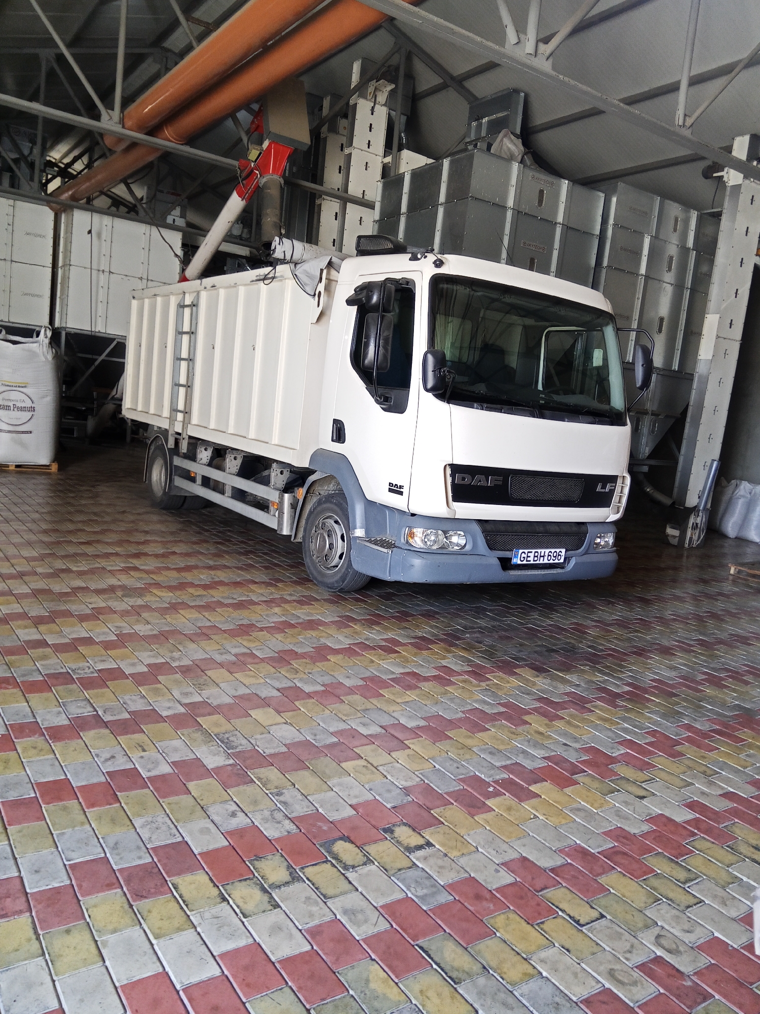 In Ceadîr-Lunga, Moldova, a white DAF LF truck is parked inside an industrial building. The truck's cargo area is enclosed and appears to be a tipper or a bulk carrier. The building features exposed metal beams and large, modular storage units stacked vertically. Orange pipes are visible overhead, suggesting an industrial process such as grain handling. The floor is paved with interlocking bricks in a pattern of red, yellow, and grey. A large white sack, possibly containing agricultural products, is positioned to the left of the truck. The license plate of the truck reads "GE BH 696". The scene appears to be within a facility related to agriculture or food processing.
