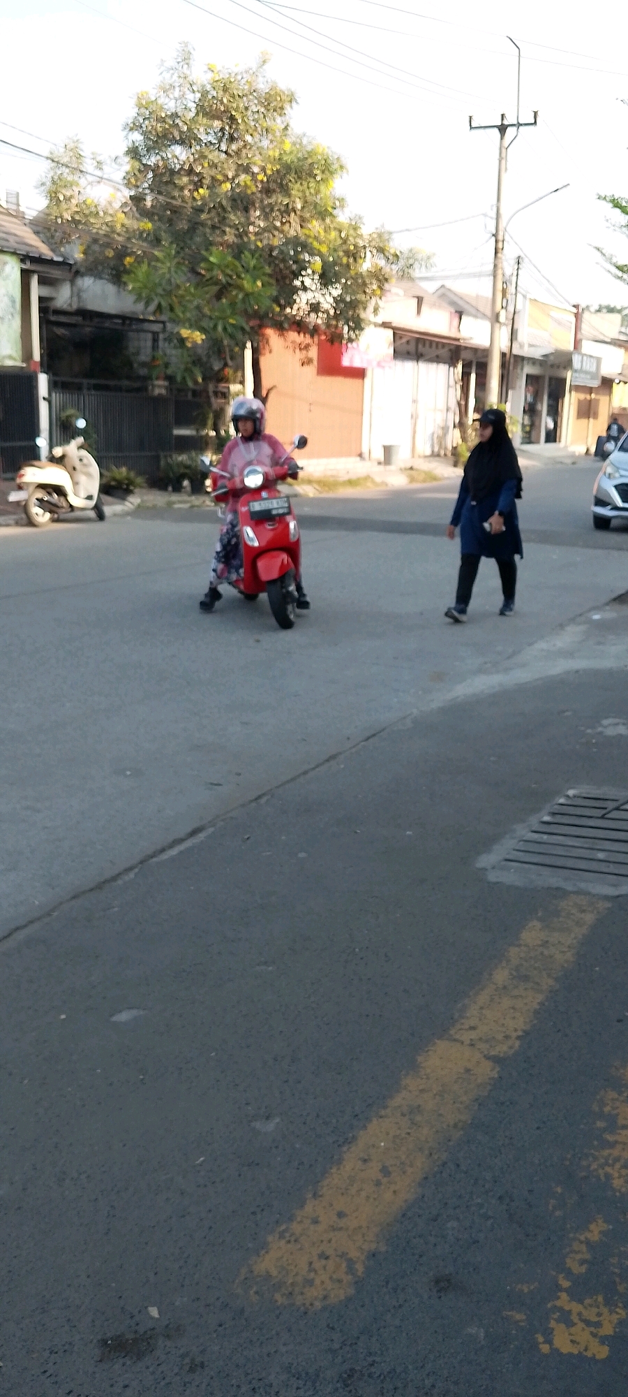 The image captures a street scene in Satriamekar, Indonesia. A person wearing a pink jacket and a helmet is riding a red scooter. To the right, another person wearing a black hijab and dark clothing is walking. In the background, there are buildings, trees, a telephone pole with wires, and a white scooter parked on the left side of the street. The road surface is asphalt with a yellow painted line.
