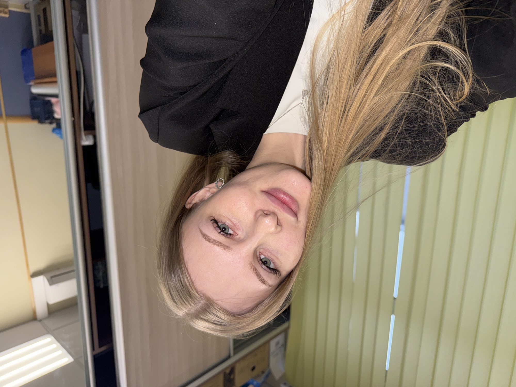 A young woman with long, blonde hair and blue-green eyes is centered in a portrait-style shot, looking directly at the camera. Her head is slightly tilted, and she has a gentle, soft expression with a faint smile. She is wearing a black blazer over a white top, and a small, circular, embellished earring is visible in her left ear.
The background suggests an indoor setting, possibly an office or a commercial building. To the viewer's left (behind the woman's right shoulder), there is a blurred area with vertical structures, appearing to be part of a cabinet or shelving unit, with faint glimpses of objects like a blue box and papers. Reflected fluorescent lighting can be seen on a surface in this area. To the viewer's right (behind the woman's left shoulder), the background features vertical, light-colored stripes, consistent with blinds or a paneled wall. The overall lighting is bright and even, indicating an indoor, artificially lit environment. The location context for this image is Perm, Russia. No visible text is present in the image.