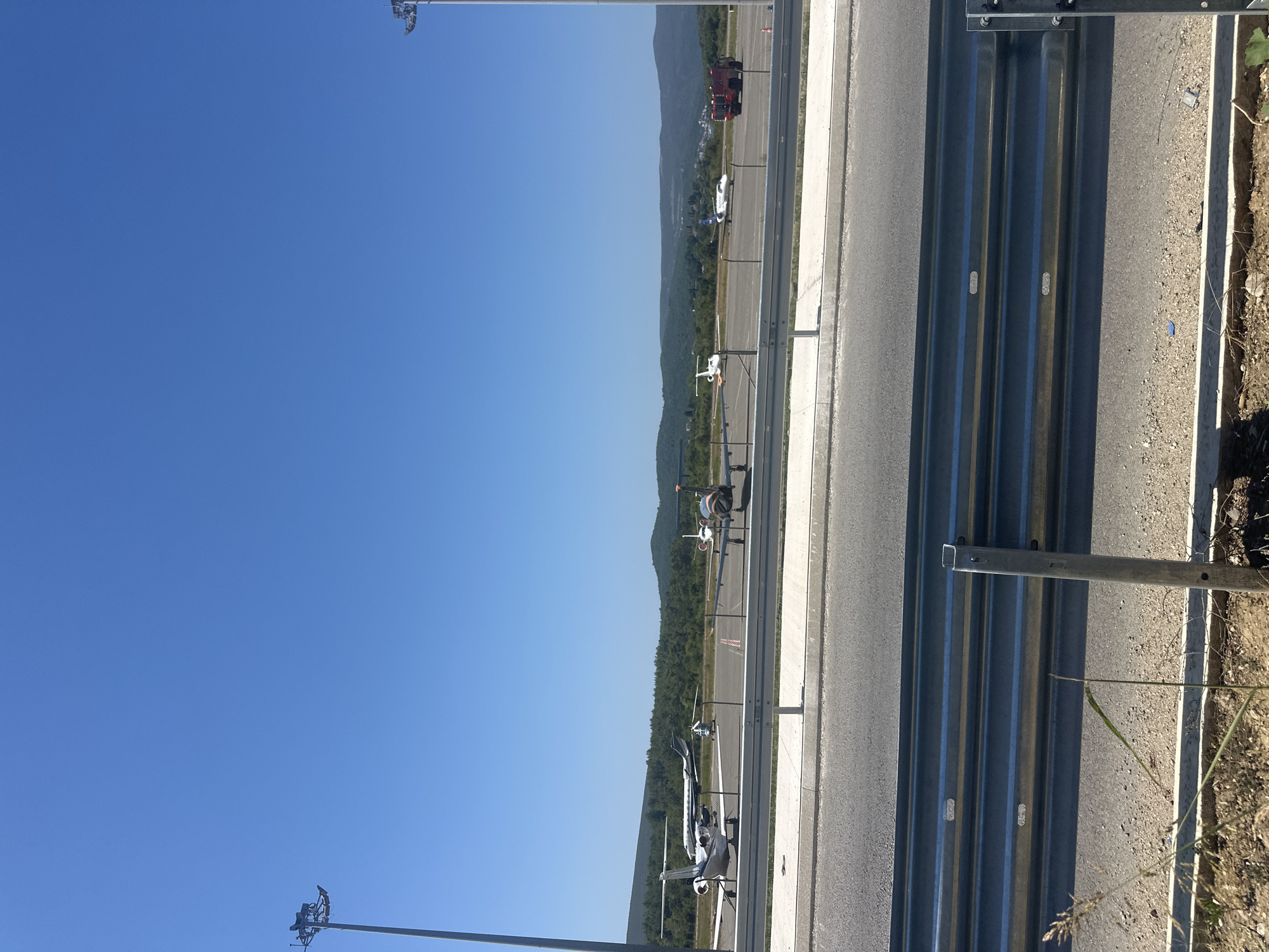 The image depicts an airfield on a clear, sunny day with a bright blue sky. In the foreground, a metal guardrail runs horizontally across the frame, with a paved surface to its left and dirt with sparse vegetation to its right. The airfield itself is visible beyond the guardrail, featuring several small to medium-sized aircraft parked on the tarmac. These include what appear to be private jets and propeller-driven planes. A chain-link fence separates the tarmac area from the foreground. In the middle ground, a line of trees forms a dense green backdrop, behind which low hills and mountains are visible. On the far right side of the airfield, a red fire truck is present. The scene is consistent with an airport environment, likely a regional or smaller general aviation airport, situated in a location with surrounding natural landscape. The city of Mrčevac, Montenegro, is a known location with an airport, Tivat Airport (LYTV), which could potentially be the setting for this image given the mountainous coastal terrain often associated with the region.