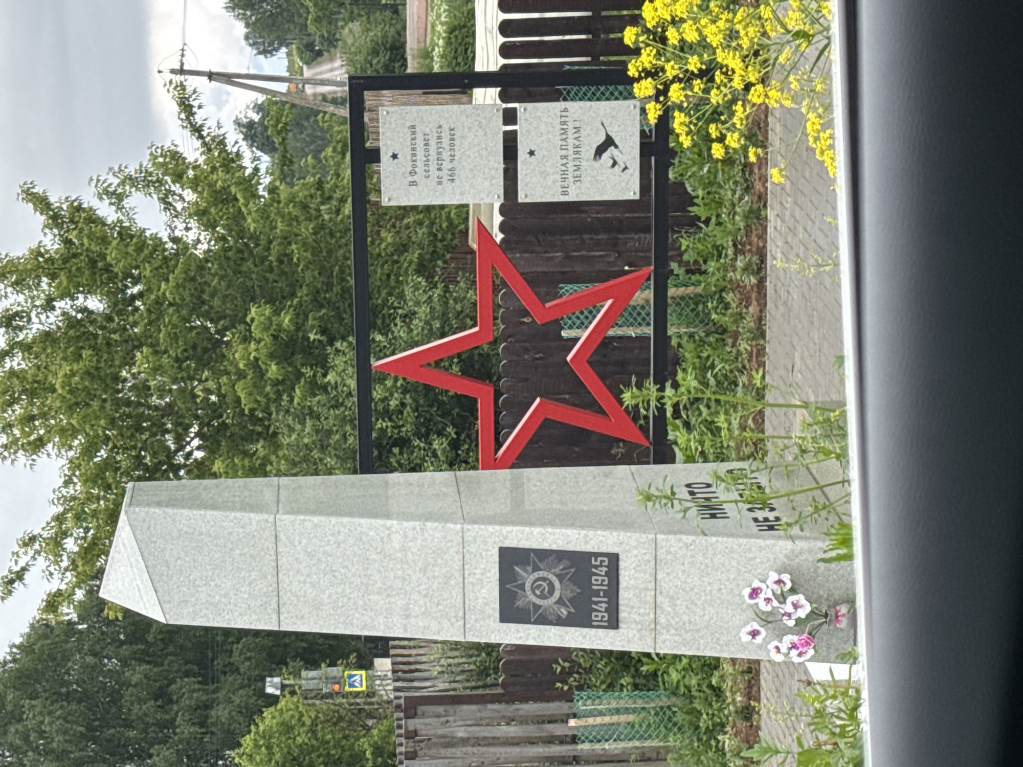 This image captures a war memorial located in the area of the Fokinsky village council, Russia. The central feature is a grey, granite-like obelisk with a sloped top. A dark rectangular plaque on the obelisk displays a Soviet Order of the Patriotic War emblem (a red star with a hammer and sickle, encircled by laurel wreaths) above the dates "1941-1945." Partially visible Cyrillic text below the plaque reads "НИКТО НЕ ЗАБЫТО" (Nobody is forgotten). At the base of the obelisk, a small vase holds a bouquet of artificial pink and white orchids.
Behind the obelisk, a large red five-pointed star is mounted within a black metal frame. To the right of the star, two white rectangular plaques are affixed to the frame. The upper plaque states in Cyrillic: "В Фокинский сельсовет не вернулись 466 человек" (466 people did not return to the Fokinsky village council). The lower plaque reads: "ВЕЧНАЯ ПАМЯТЬ ЗЕМЛЯКАМ!" (Eternal memory to our countrymen!) and features a silhouette of two flying birds.
The background includes a wooden fence, followed by abundant green trees and foliage under an overcast sky. A dirt road with utility poles and power lines extends into the distance on the right. To the left, a partial view of a traffic light and a blue pedestrian crossing sign are visible. The immediate foreground on the right features a cluster of bright yellow flowering plants. The bottom edge of the image shows a dark grey car interior element, suggesting the photo was taken from a vehicle. No individuals are present in the scene.
