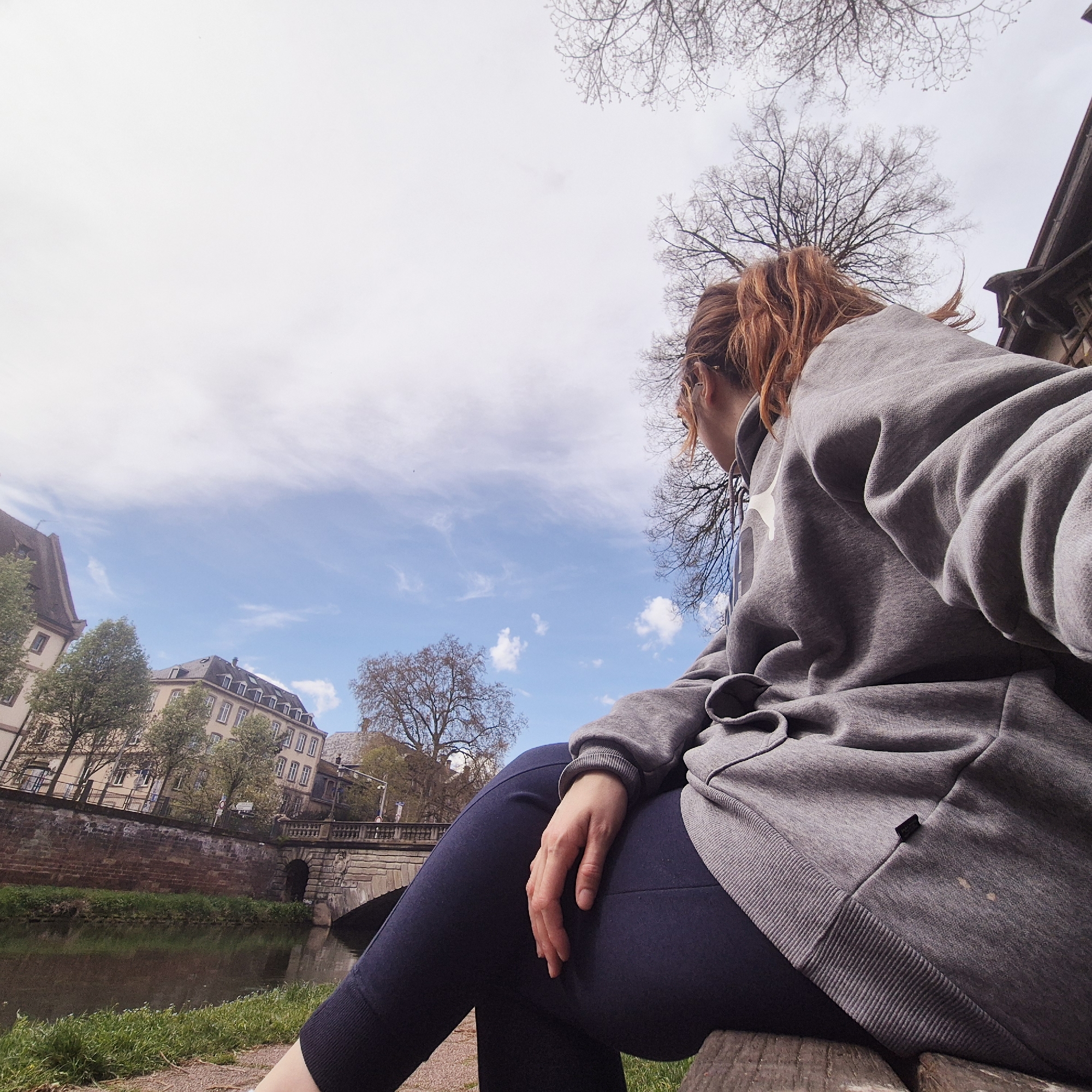 A woman with red hair sits on a bench near the Ill River in Strasbourg, France. She wears a grey hooded sweatshirt and dark pants. In the background, there are buildings, bare trees, and a small stone bridge over the river. The sky is light blue with some clouds. The image appears to be taken from a low angle.