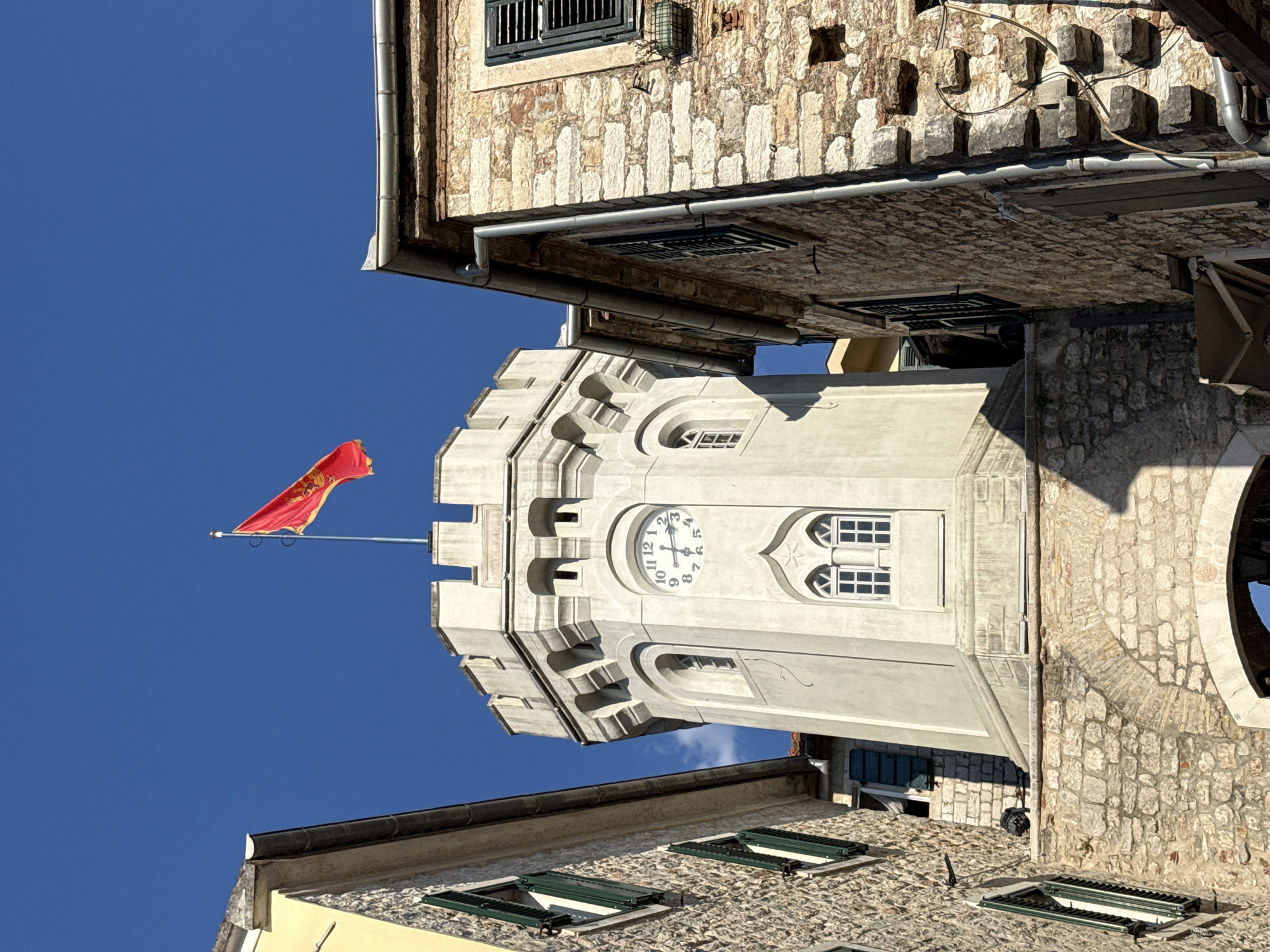A low-angle, full shot captures a tall, octagonal stone clock tower with a red and gold flag flying from its peak, set against a vibrant, cloudless blue sky. The tower, which appears to be part of a historic building, is made of light-colored stone and features crenellated battlements and arched windows. A circular clock face with Roman numerals is prominently displayed on one side of the tower. The tower is flanked by two older buildings constructed from rough, reddish-brown stone, with dark shutters on their windows. The building on the left has a yellow facade and a light-colored roofline. The building on the right has a more weathered appearance with some stones missing. A stone archway is visible at the base of the tower, leading into a shaded area. This scene is likely the main entrance of the Forte Mare fortress in Herceg Novi, Montenegro.
