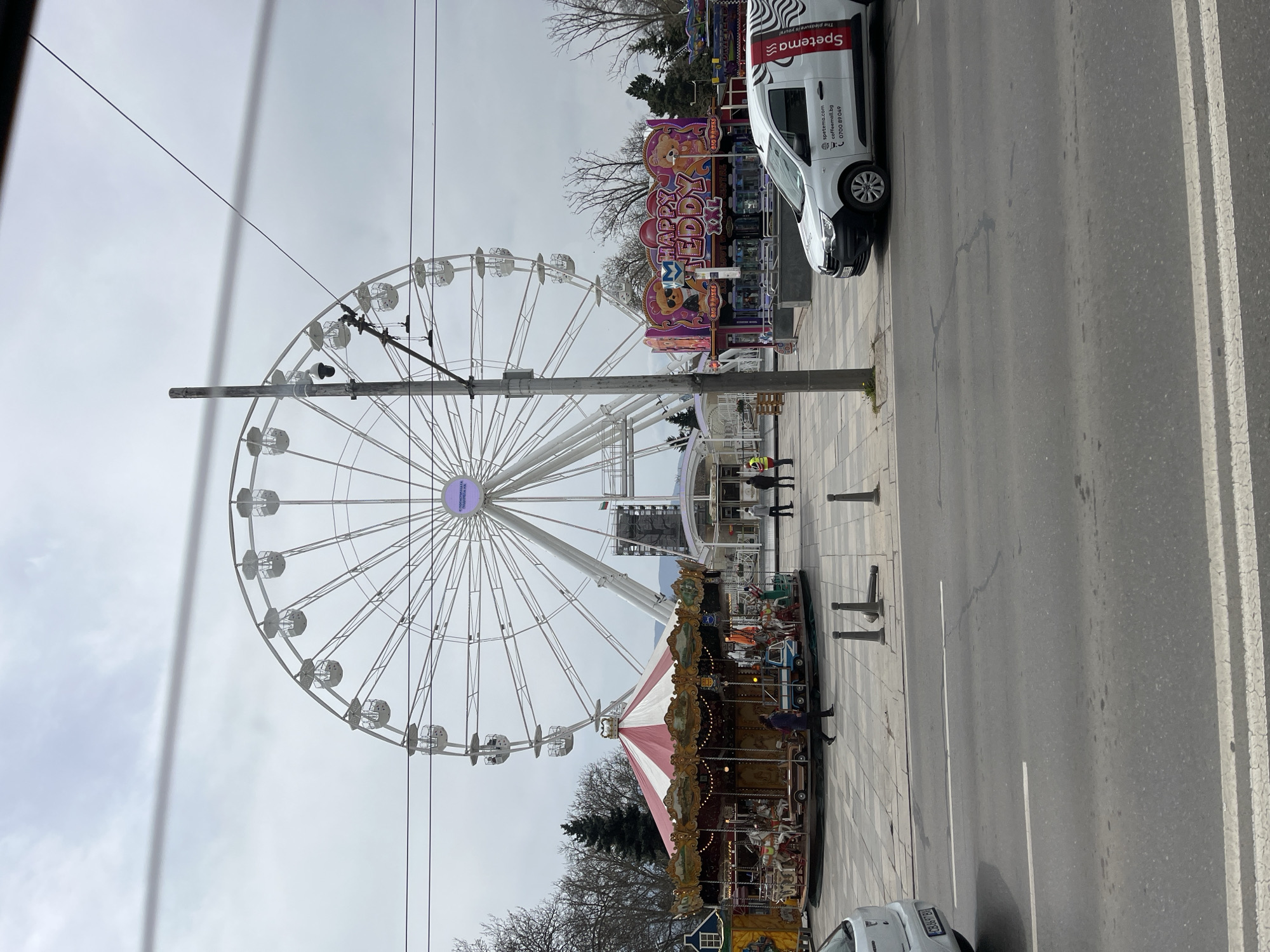 The image depicts a Ferris wheel and carousel located in the city of Unknown, Unknown, on a street parallel to a multi-lane road. A white Spetema van is parked nearby. A tall utility pole is situated between the Ferris wheel and the road, with power lines strung across the sky. The weather appears overcast.