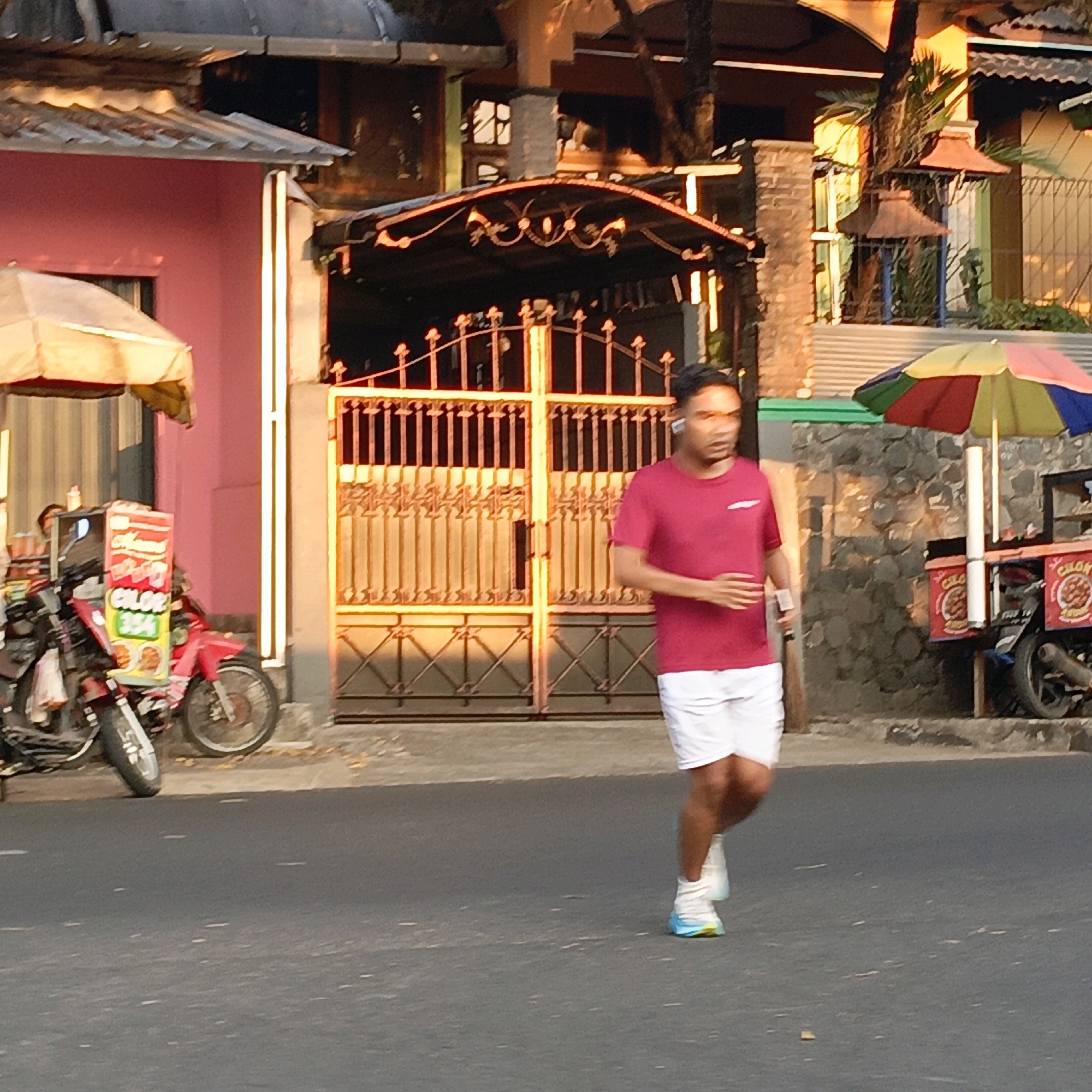 The image depicts a man jogging on a paved street in Salatiga, Indonesia. He is wearing a red t-shirt and white shorts, and athletic shoes with blue and yellow accents. The man appears to be wearing wireless earbuds. Behind him, a tall, ornate metal gate, likely the entrance to a property, stands open. To the left of the gate, a pink building with a corrugated metal roof is visible, partially obscured by an awning. A parked motorcycle with a food vendor cart attached to it is situated near the pink building. The cart is decorated with red and white signage featuring food imagery and text in Indonesian. To the right of the gate, a multi-colored umbrella, red, yellow, green, and blue, is open and attached to another food vendor cart, which is also mounted on a motorcycle. This second cart displays round signs with food illustrations and text in Indonesian. The ground is asphalt, with some scattered leaves. The lighting suggests it is either early morning or late afternoon, with the warm tones indicating sunlight.
