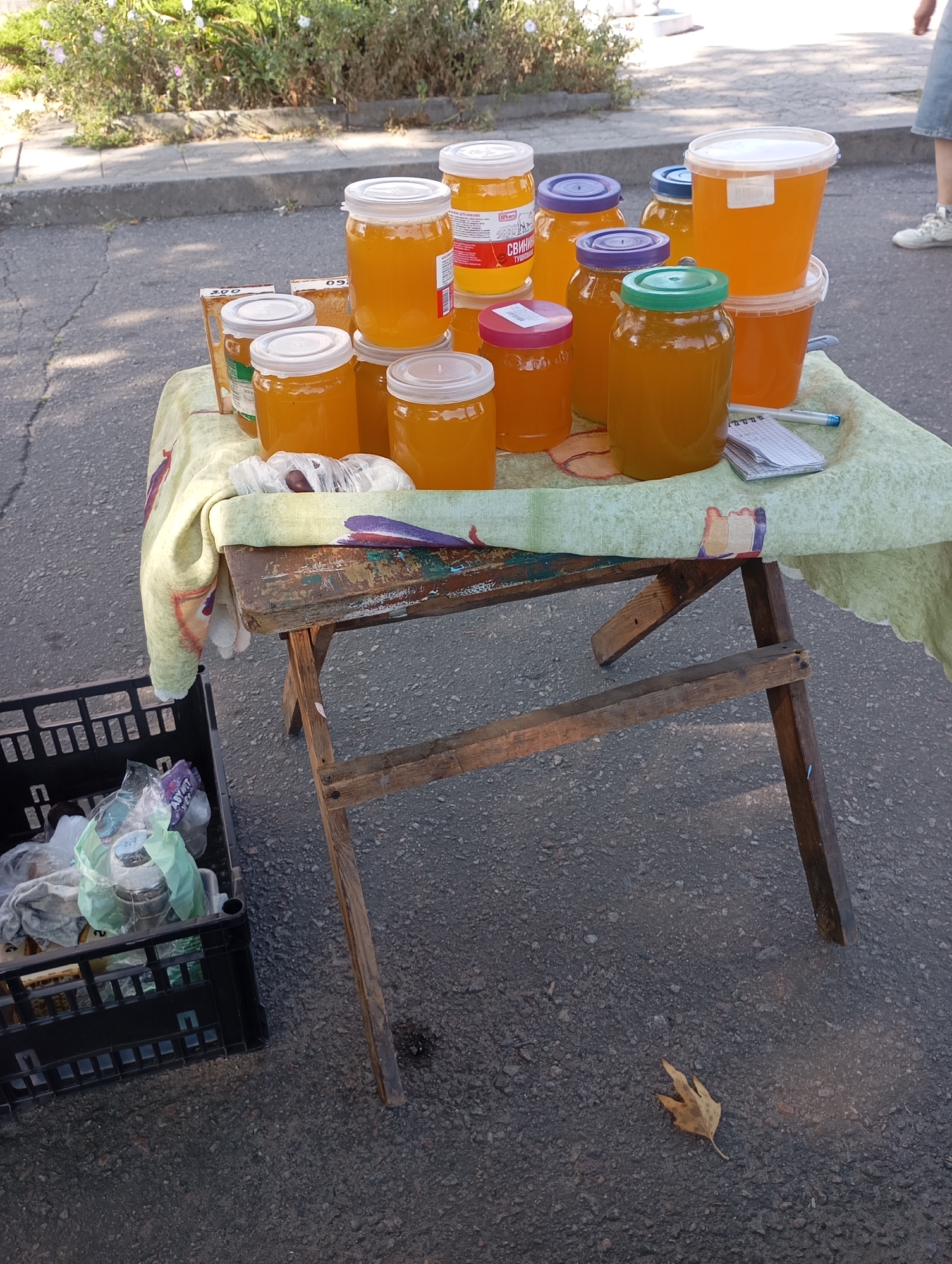 A sidewalk market stall is set up on a paved street in Horishni Plavni, Ukraine. The stall consists of a weathered wooden table covered with a light green cloth patterned with colorful illustrations. Displayed on the table are several glass jars and plastic containers filled with golden honey. Some jars have white lids, while others have blue or green lids. A small notepad and a pen are also present on the table. To the left of the table, a black plastic crate is filled with various items, including plastic bags containing what appear to be groceries. In the background, lush green foliage and some flowering plants are visible. The ground surface is asphalt, and a fallen brown leaf rests near the table leg. The overall scene depicts a street vendor selling honey.